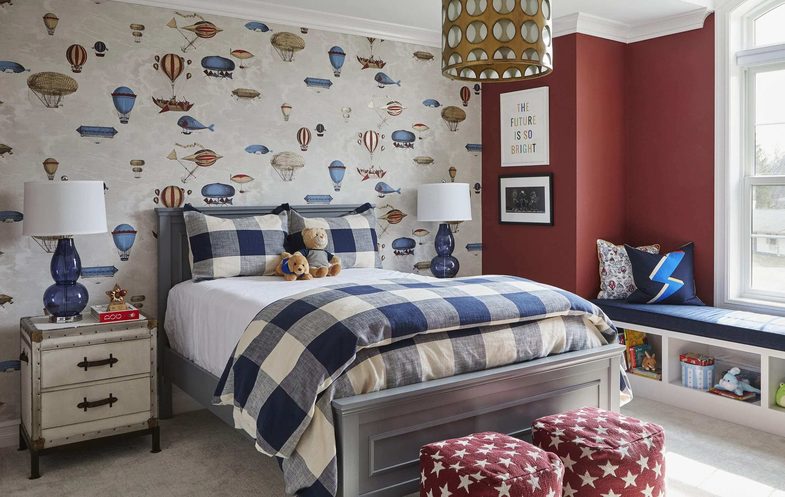 A bedroom with a twin bed, blue and white plaid bedding, and two teddy bears. The wall behind has wallpaper with hot air balloons and airships. There are side tables with lamps, framed pictures, and a window with seating and books. The room has a red