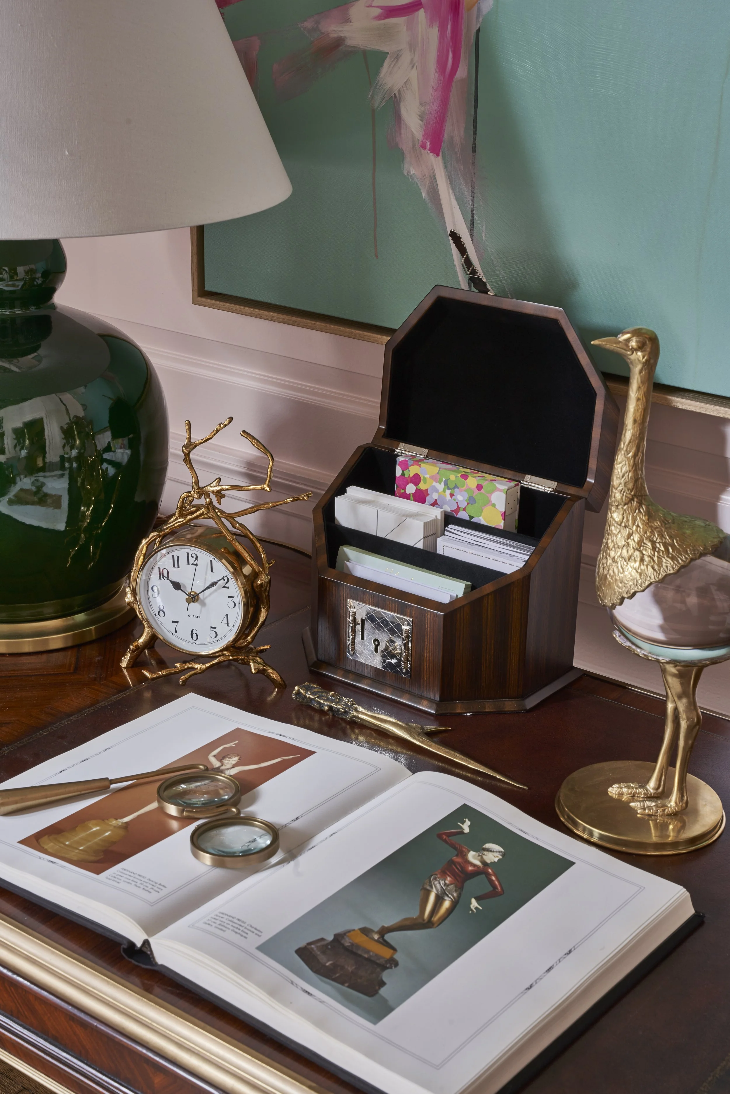 Decorative desk items including a large golden goose sculpture, a vintage clock, a wooden jewelry box, and open fashion books with jewelry and illustrations on a wooden table, with a framed abstract painting in the background.