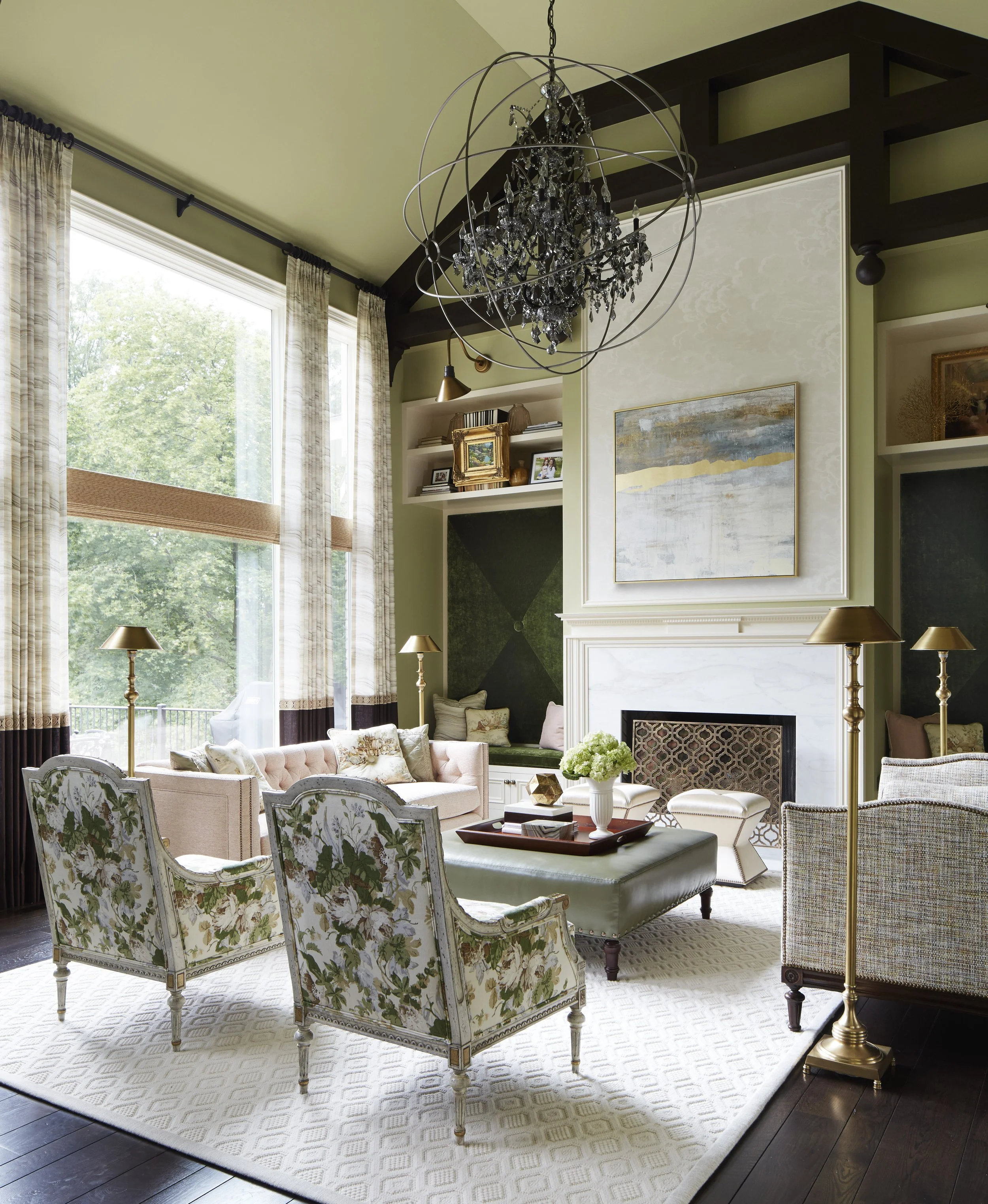 A living room with large windows, beige curtains, and a mix of elegant furniture including floral armchairs, a pink tufted sofa, lamps, and wall art. A chandelier hangs from the ceiling.