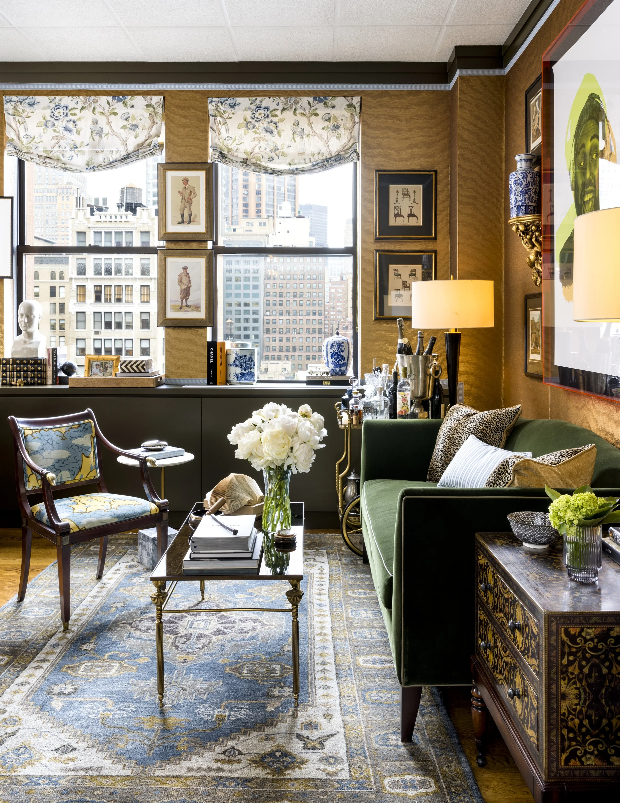 Interior of a stylish living room with a large window showing city buildings, decorated with framed art, vases, a table with books, and a green sofa with patterned pillows. The room has a patterned rug and elegant furniture.