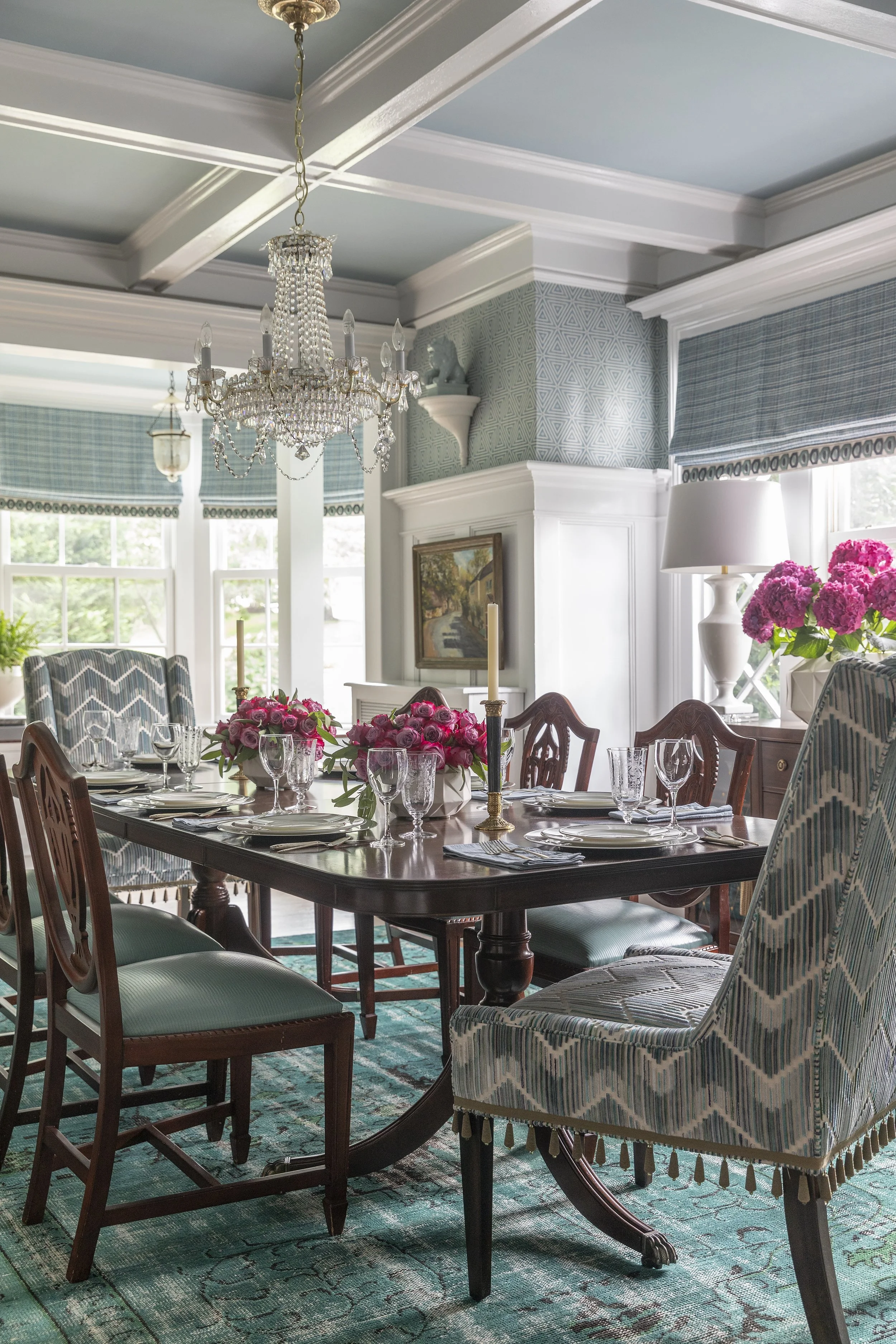 A formal dining room with a large wooden dining table set with pink roses and white candles, surrounded by patterned and solid-color chairs, with a chandelier hanging from the ceiling and large windows with blue patterned curtains, a sideboard with a