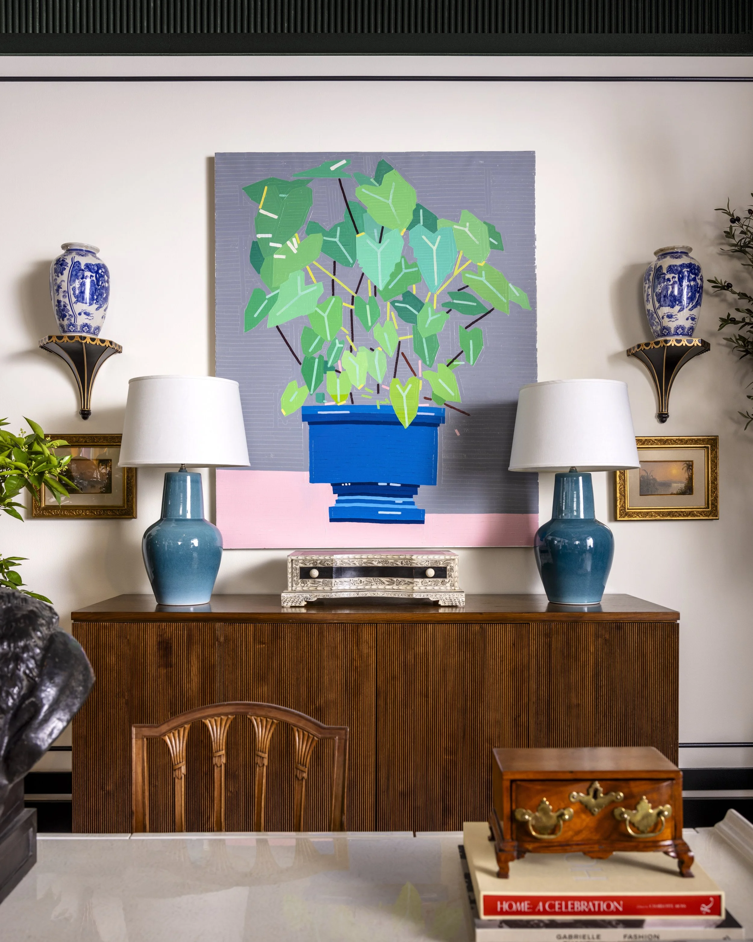 Interior of a home with decorative items. There are two blue and white vases on black and gold wall sconces on either side of a large painting of a green potted plant. Below the painting are two white lamps. In front, a wooden table holds a small orn