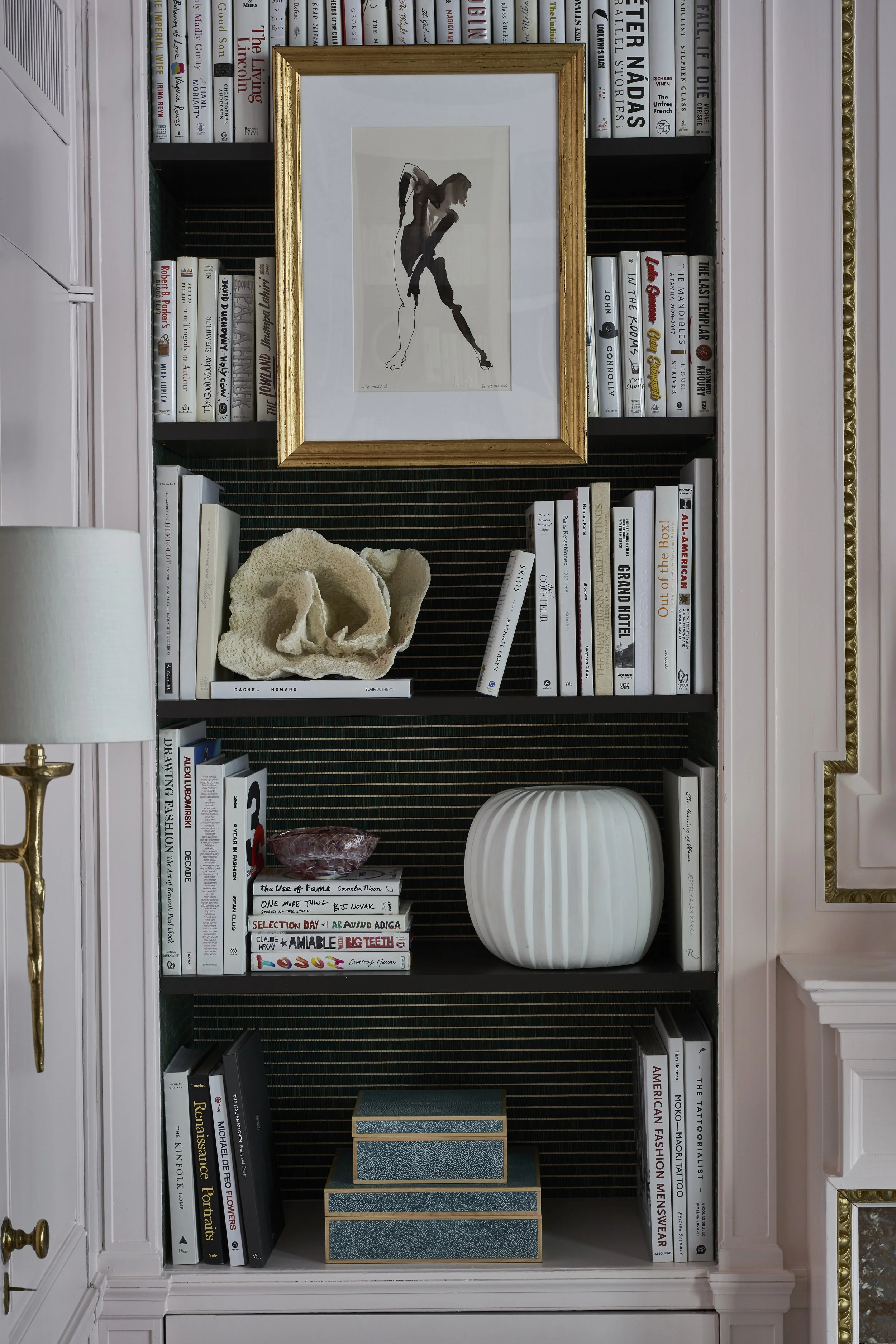 A bookshelf with two top shelves filled with books, a middle shelf with a decorative coral sculpture and books, and a bottom shelf with books and a stacked display box. There is a framed artwork of a woman playing tennis on the wall behind the booksh
