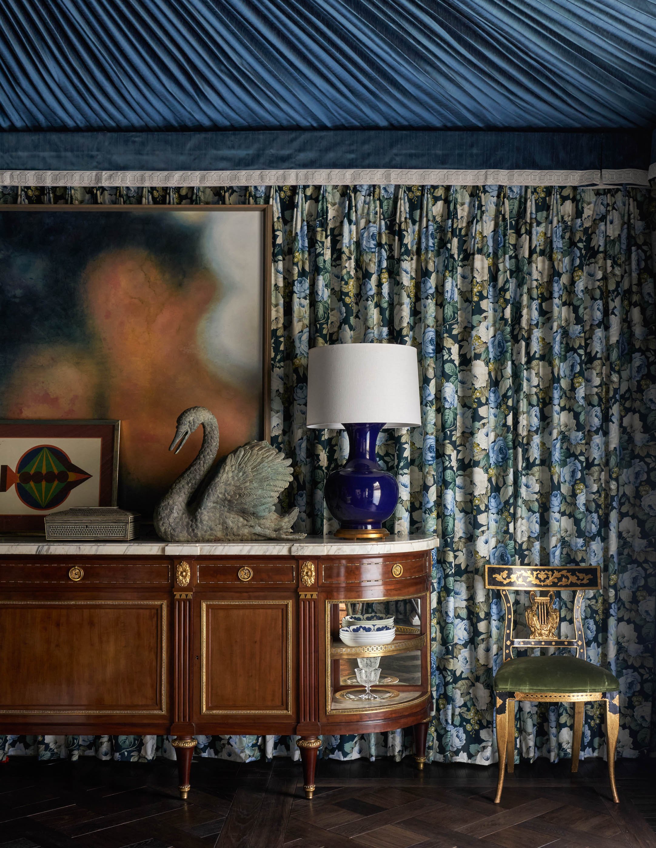 An elegant room interior featuring a large wooden sideboard with decorative gold accents, a blue and white ceramic bowl on a small side table, a white lampshade on a blue ceramic base lamp, a gold and black chair, and a black velvet stool. The backgr