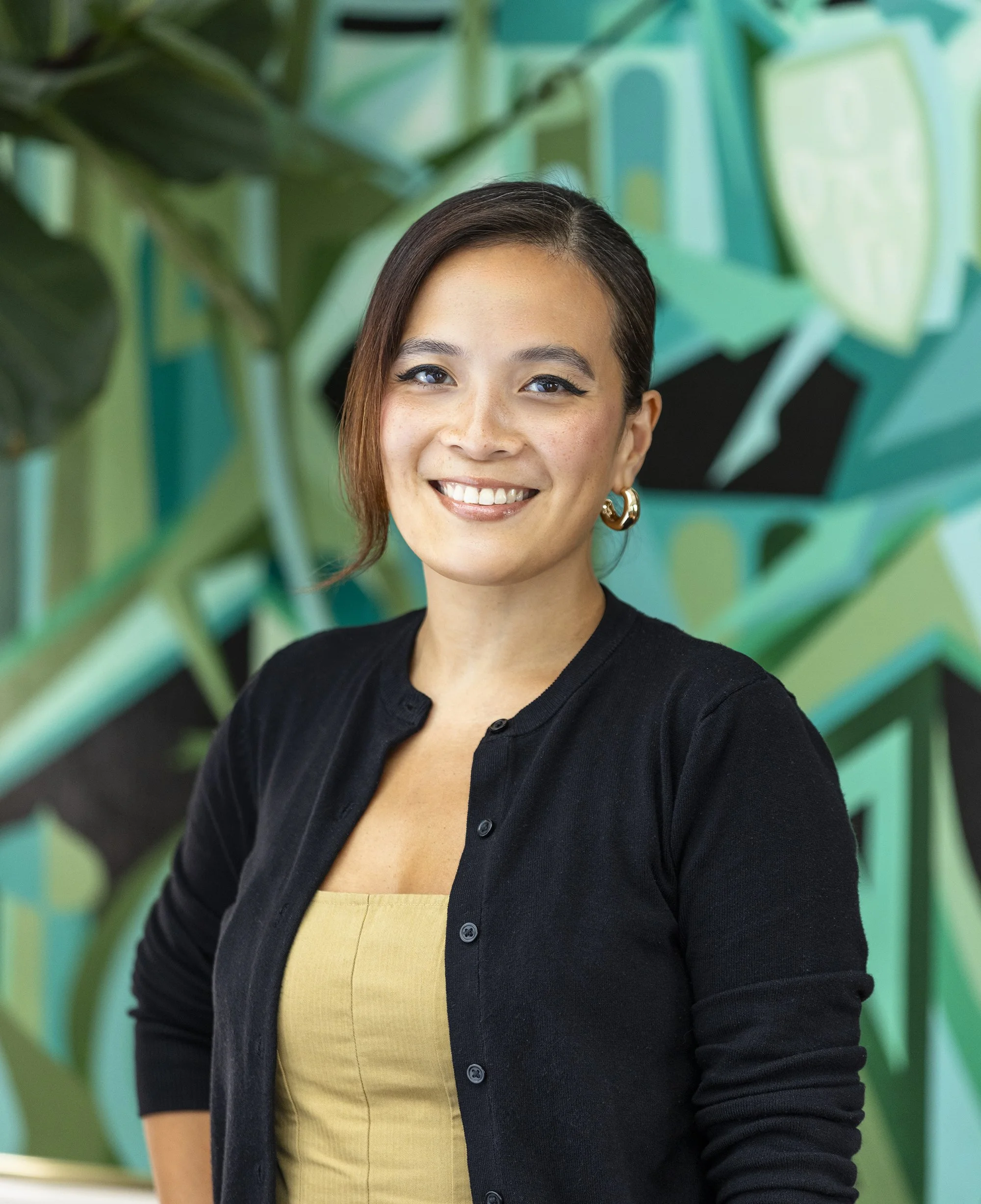 A young woman with short brown hair, blue eyes, and hoop earrings smiling in front of a colorful, abstract green and blue background. She is wearing a black cardigan over a yellow top.