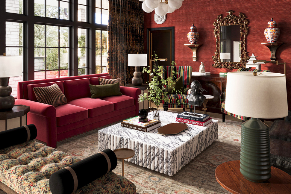 A living room with a large red velvet sofa, striped armchairs, and black cylindrical pillows. A marble coffee table holds books and decorative items. The room features large windows with dark curtains, a red textured wall with an ornate gold-framed m