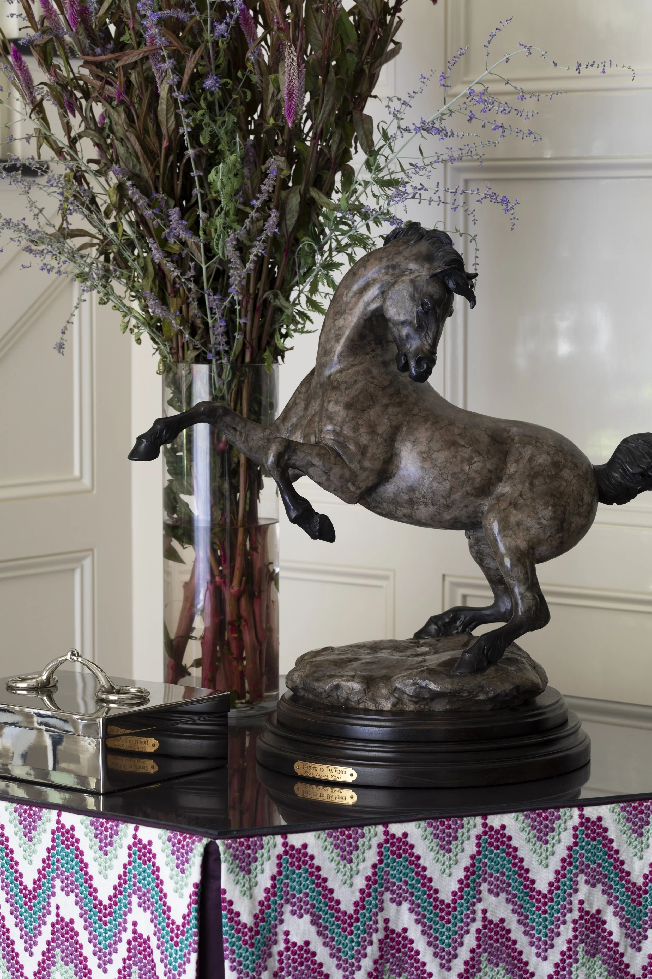 A bronze horse statue on a black marble base on a table, with a large glass vase of purple and green foliage in the background, and a silver decorative box on the table. The table is covered with a patterned cloth with pink, purple, and teal designs.