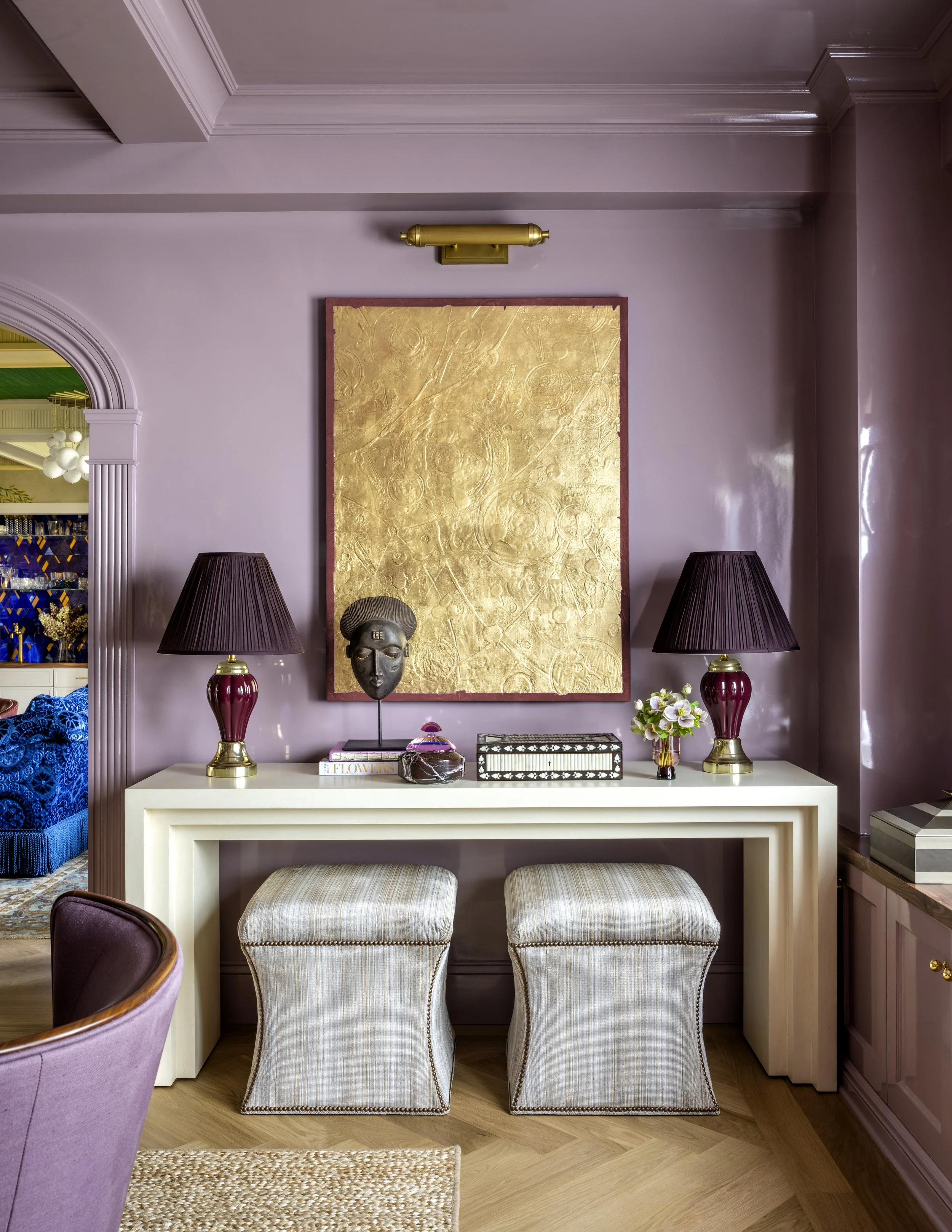 A living room setup with a white console table, two upholstered stools, a decorative mask, a plant, and black table lamps, with a gold abstract artwork on a lavender-colored wall.