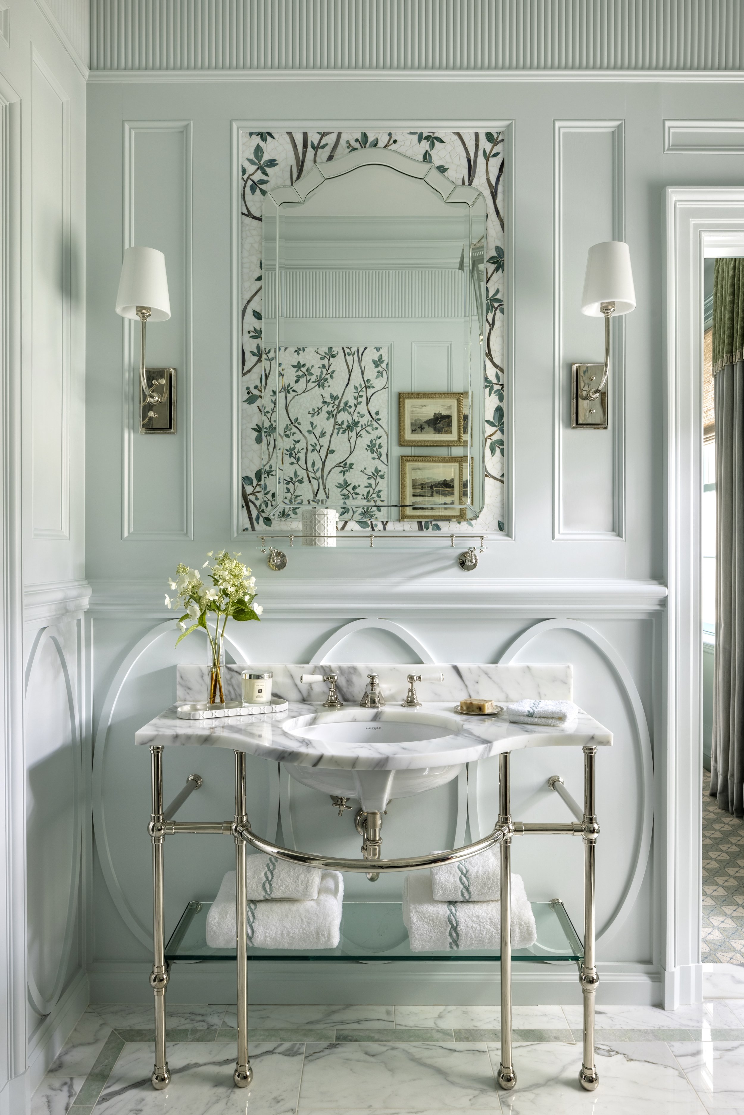A luxurious bathroom with a white marble sink, a large mirror, and wall-mounted lamps. A towel and decorative items are on the sink, and a towel rack holds two neatly folded towels. The decor is elegant with greenery accents and framed artwork visibl