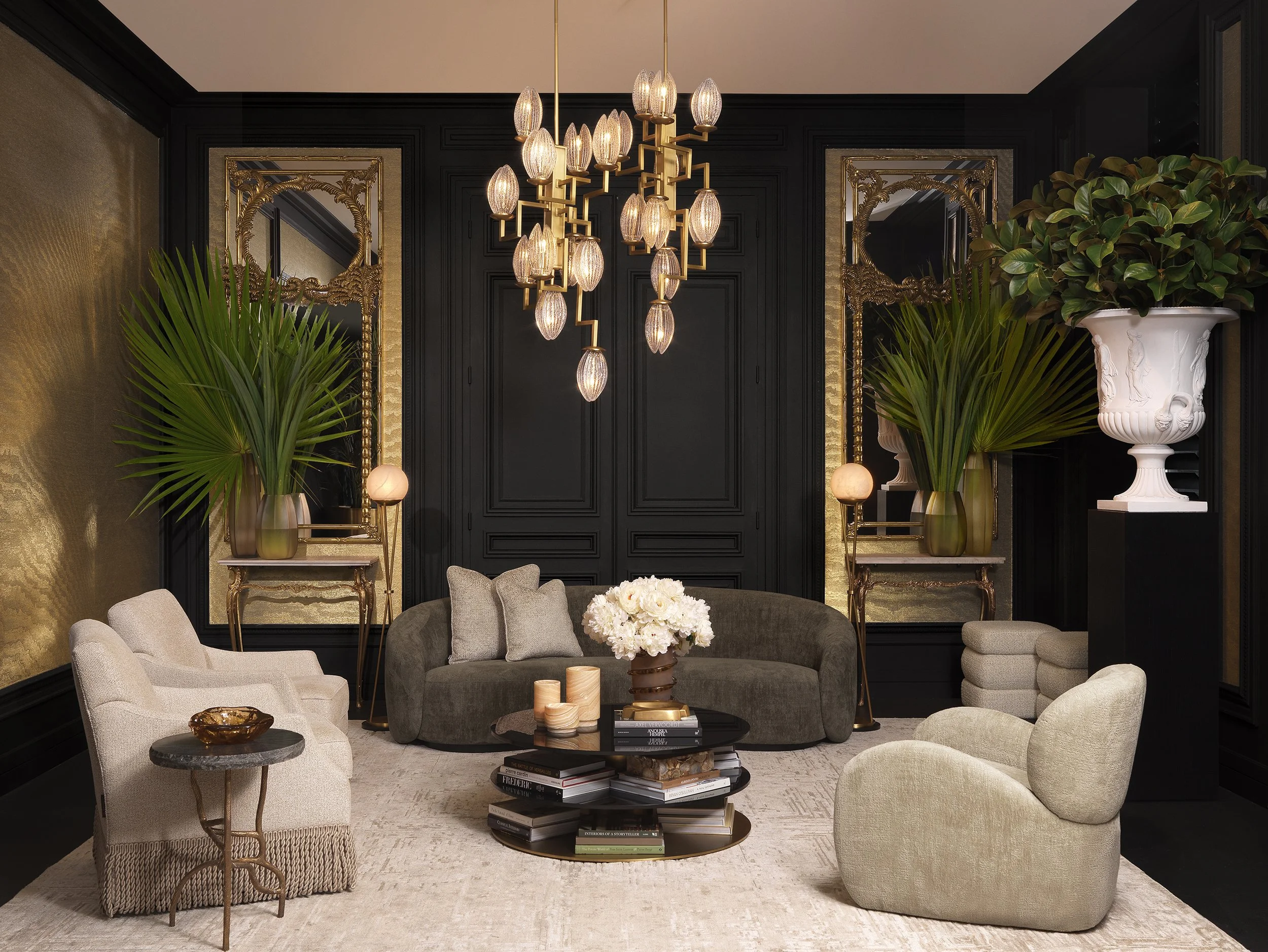 Elegant living room with black paneled wall, gold accents, a modern chandelier with multiple glass bulbs, a round gray sofa, cream-colored armchairs, a black coffee table with stacked books and decorative items, large green plants in white and gold p
