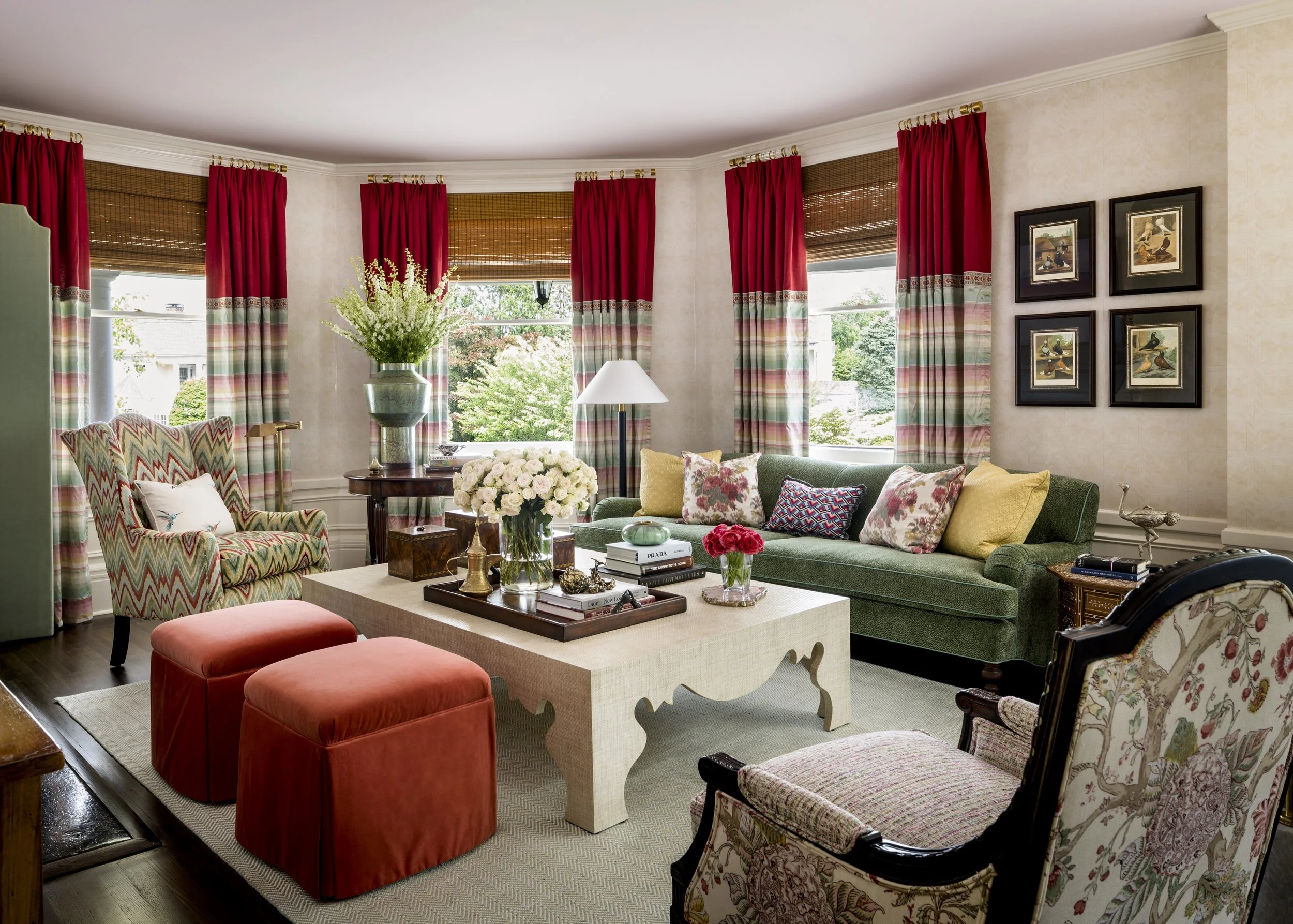 Living room with large windows, red curtains, floral cushions, a green sofa, floral armchair, and a white coffee table with books and flowers.