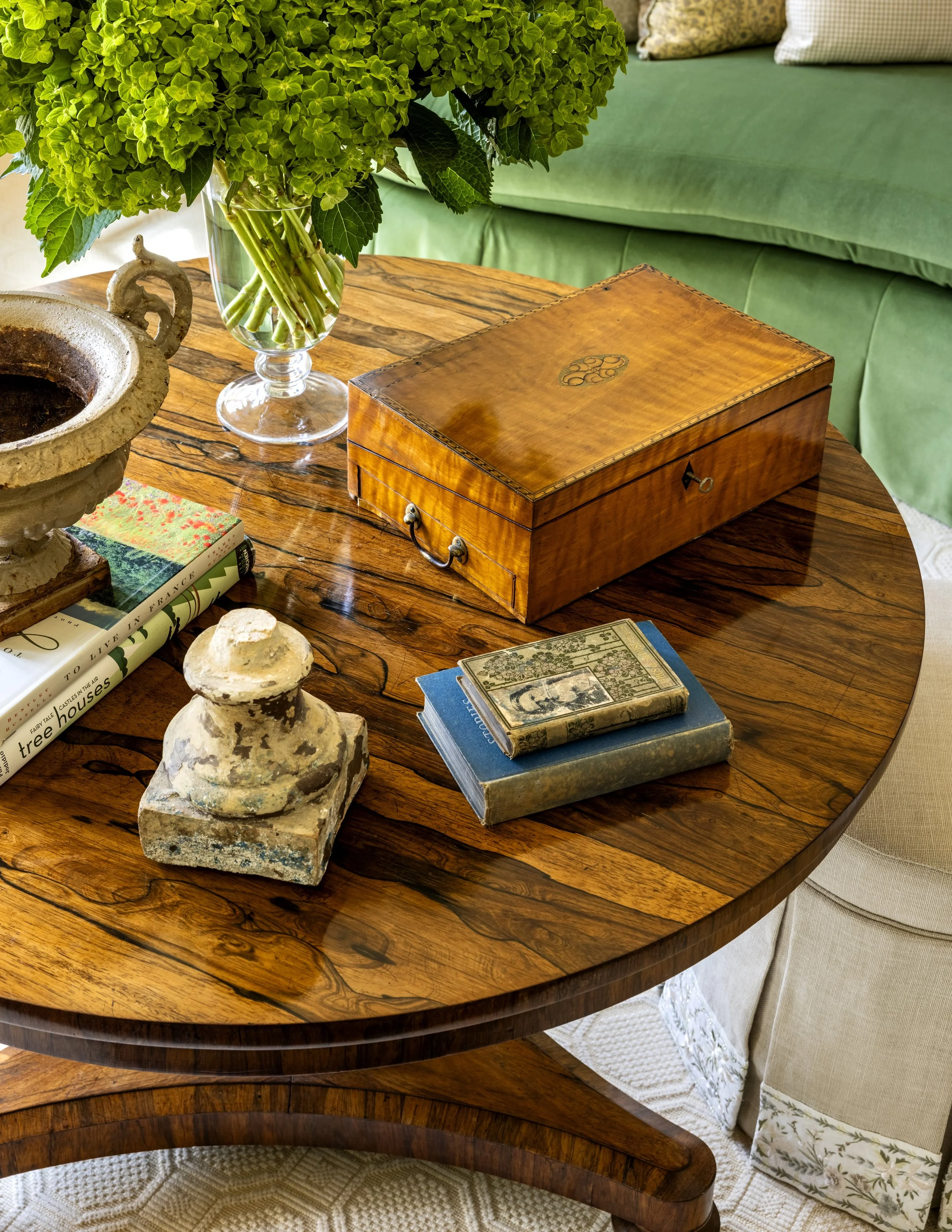 A wooden table with a green sofa in the background. The table has a glass vase with green hydrangea flowers, a decorative stone urn, two nearby books, a wooden jewelry box, and a vintage-style book. Part of a cream-colored armchair and patterned pill