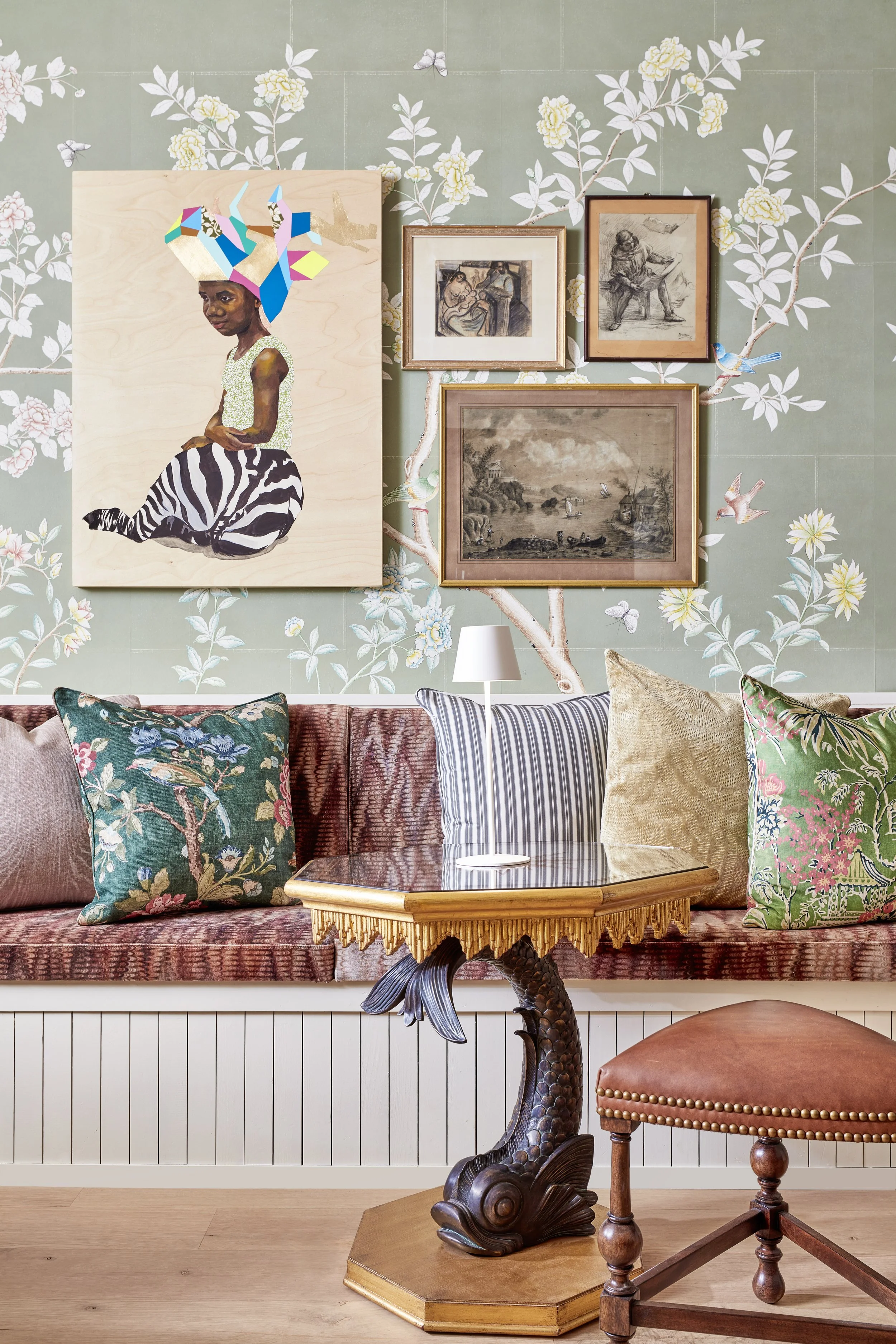 Interior of a living room with floral wallpaper, framed artwork, pillows with floral and nature motifs, a table with an intricate carved base, and a brown upholstered stool.