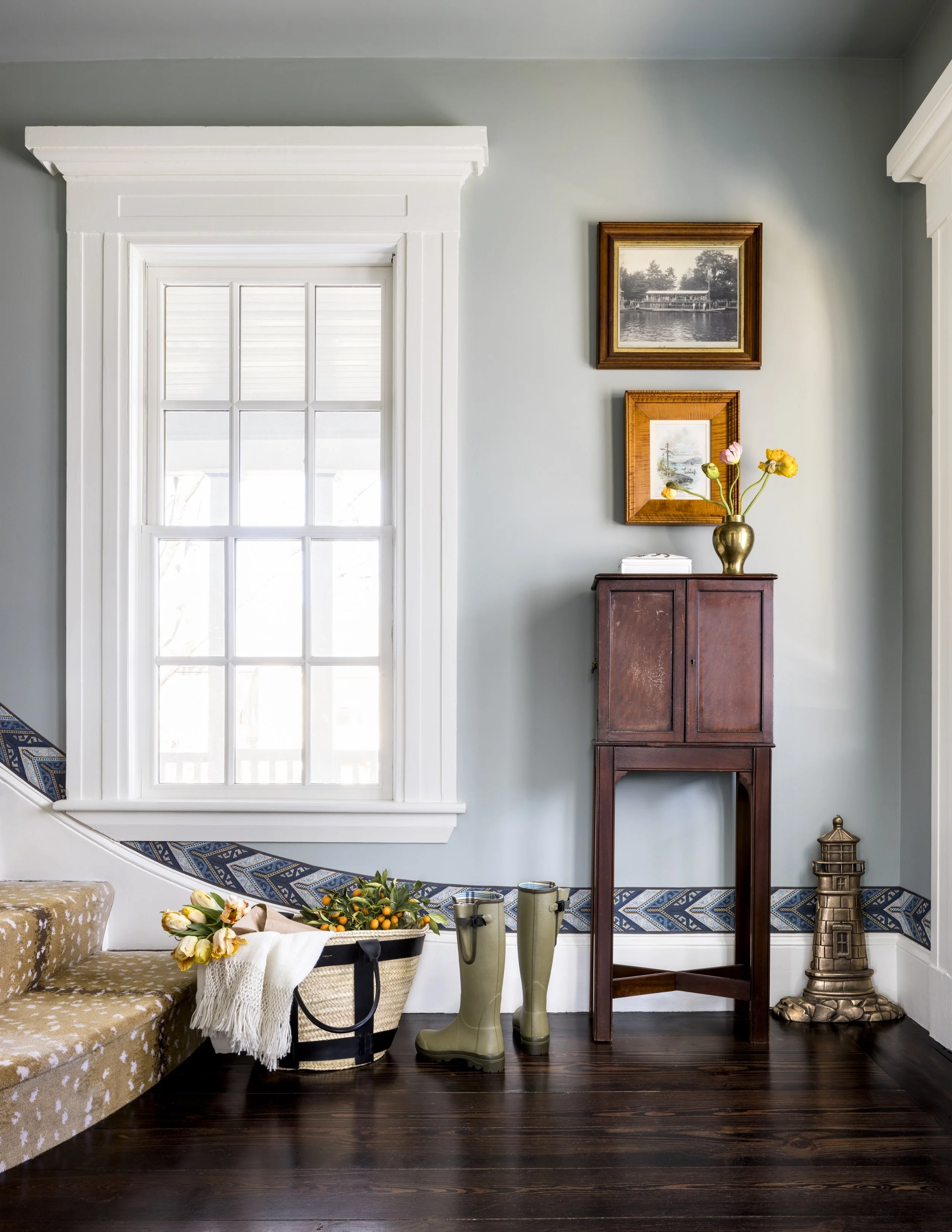 Interior of a home with a large window, a small dark wood cabinet, and decorative items including a vase with flowers, a basket with flowers, and tall rain boots. Artwork hangs on the wall, and there's a small ornate lighthouse sculpture on the floor