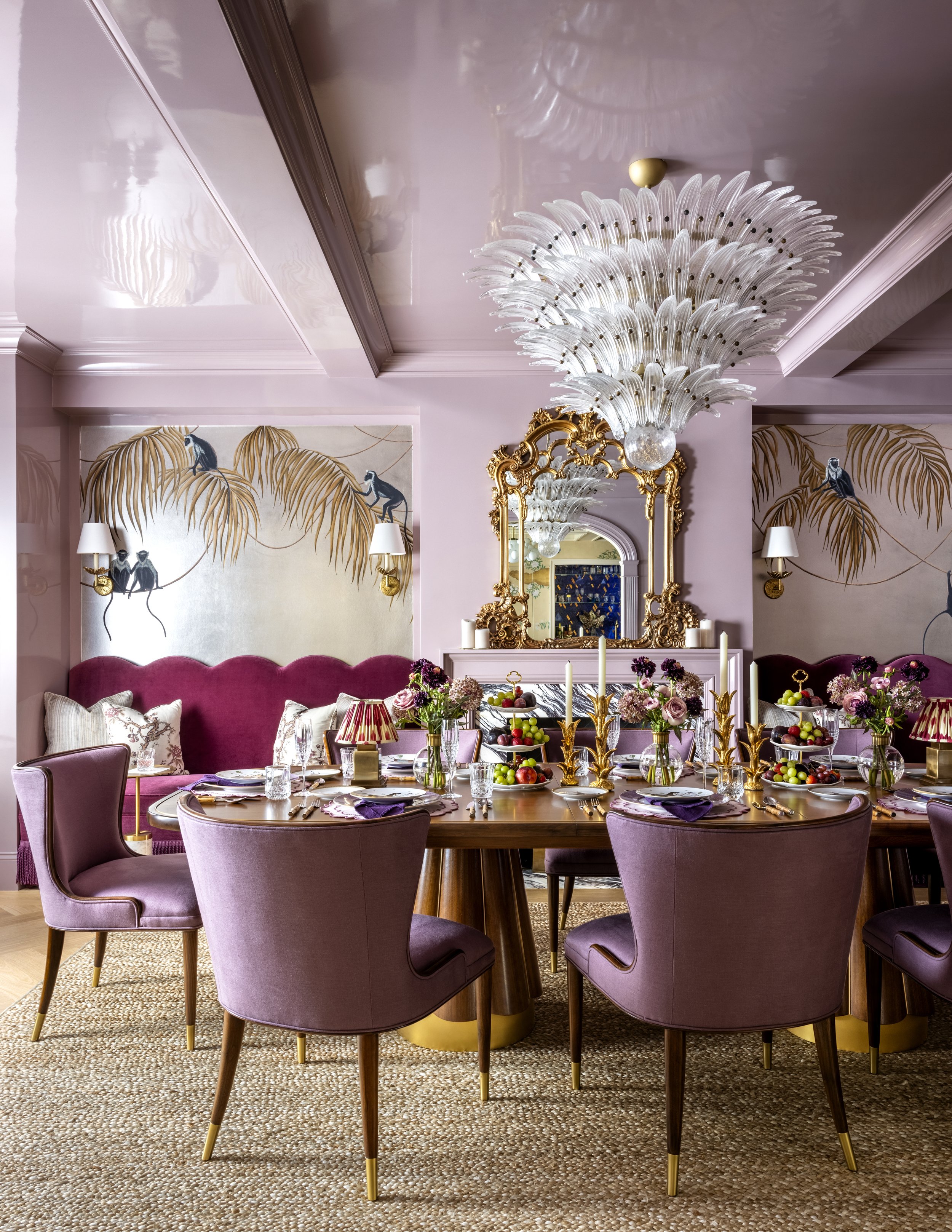 Elegant dining room with a large purple velvet sofa, pink walls, modern artwork of monkeys and palm leaves, a crystal chandelier, a gold-framed mirror, and a table set with flowers, fruits, candles, and fine tableware.
