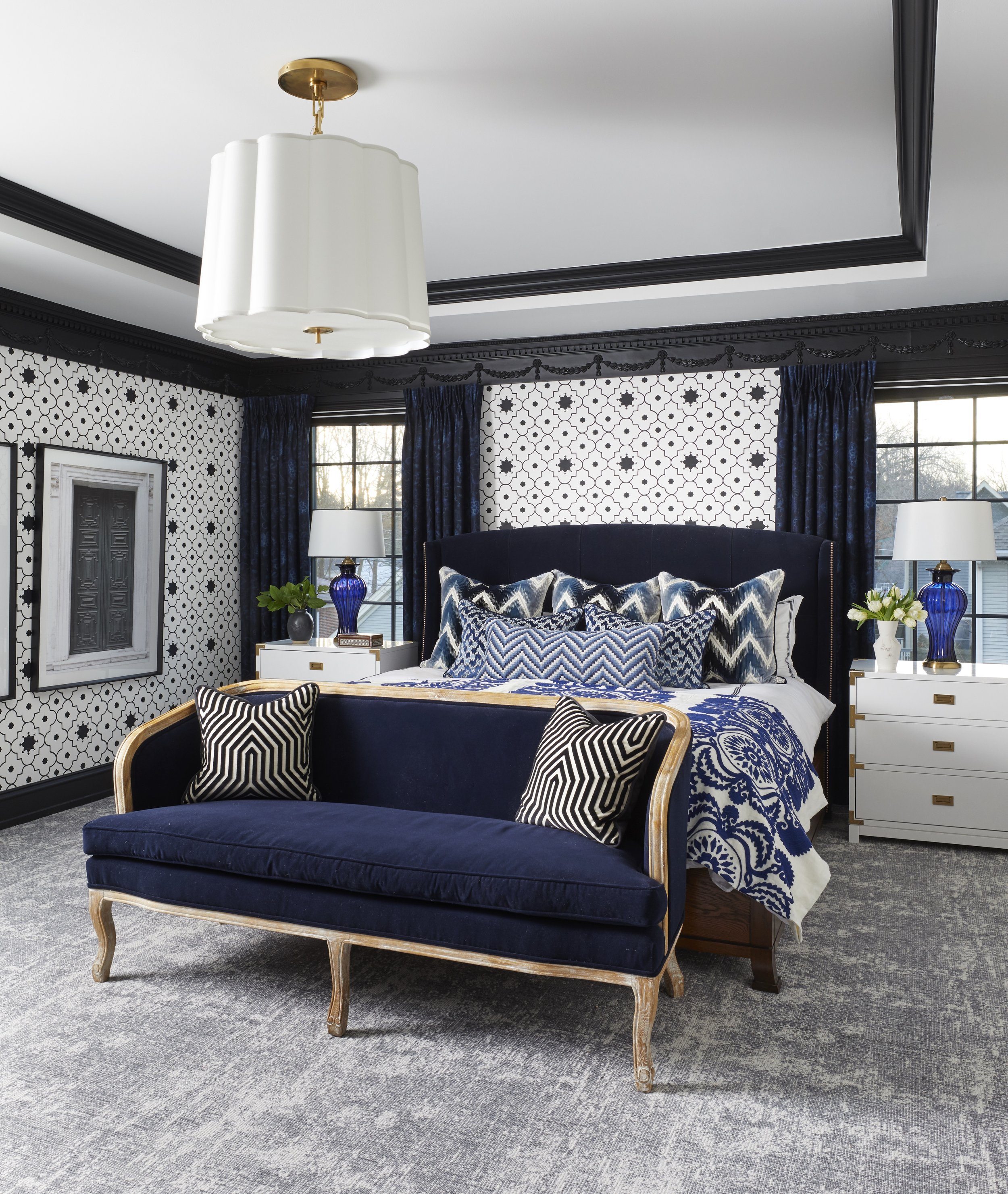 A bedroom with black and white patterned wallpaper, a navy blue velvet bed with multiple decorative pillows, a white nightstand with gold handles, a blue glass table lamp, a white lampshade, and curtains matching the wallpaper. There are large window