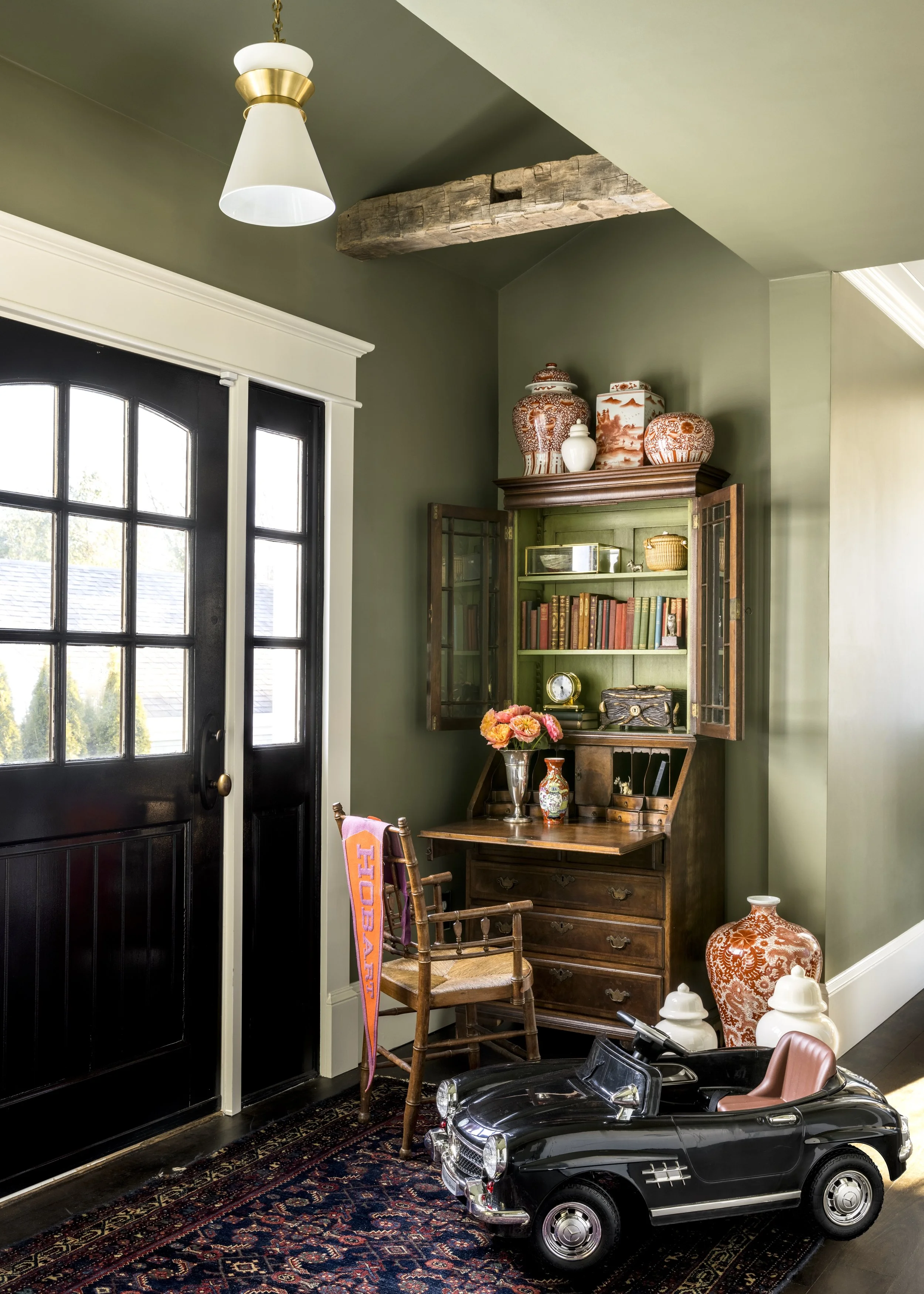 A corner of a room with dark wooden American style furniture, vintage decorative items, and a black toy Mercedes-Benz convertible car. There are vases, a floral arrangement, books, and decorative jars on a green cabinet, with a hanging ceiling light 
