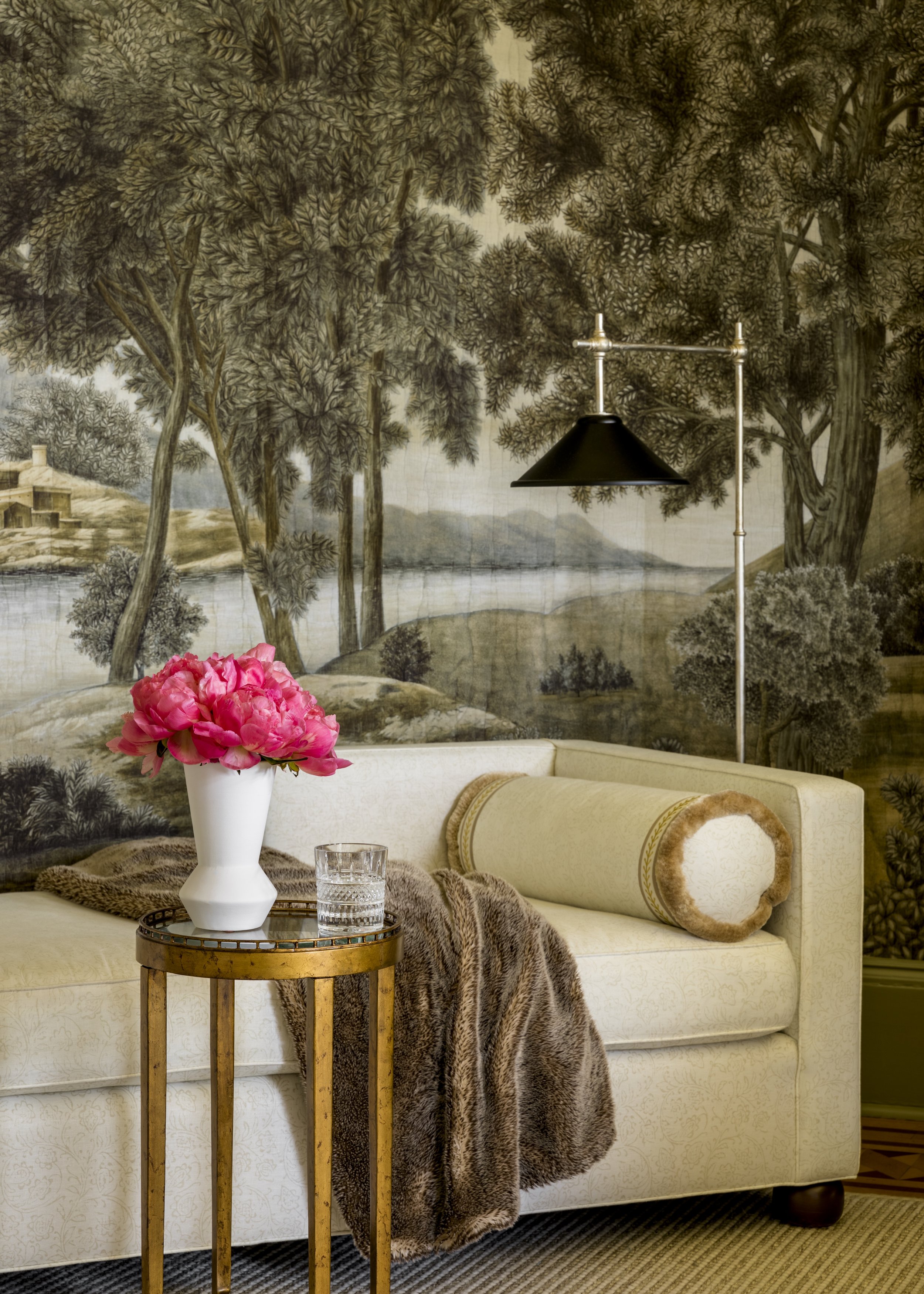 A cozy living room features a white upholstered sofa with a fur throw and decorative pillow. A small round gold side table holds a white vase with pink peonies and a glass of water. Behind, a mural of a landscape with trees and a river adorns the wal
