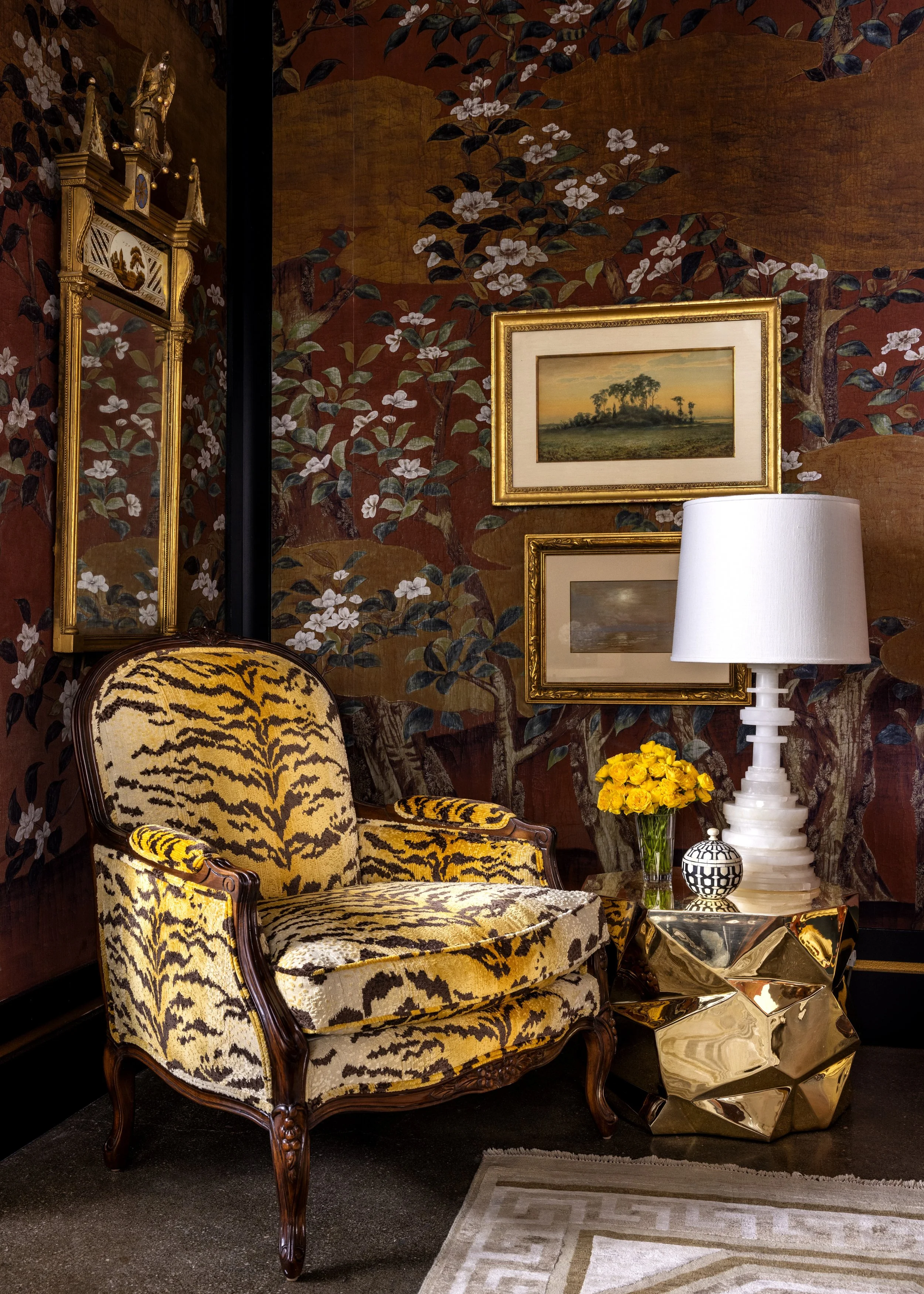 An elegant interior living space featuring a gold-framed armchair with tiger print upholstery, a matching ornate armrest, a vase with yellow flowers, a white table lamp, and framed artwork on a richly patterned wall with floral motifs. A reflective g