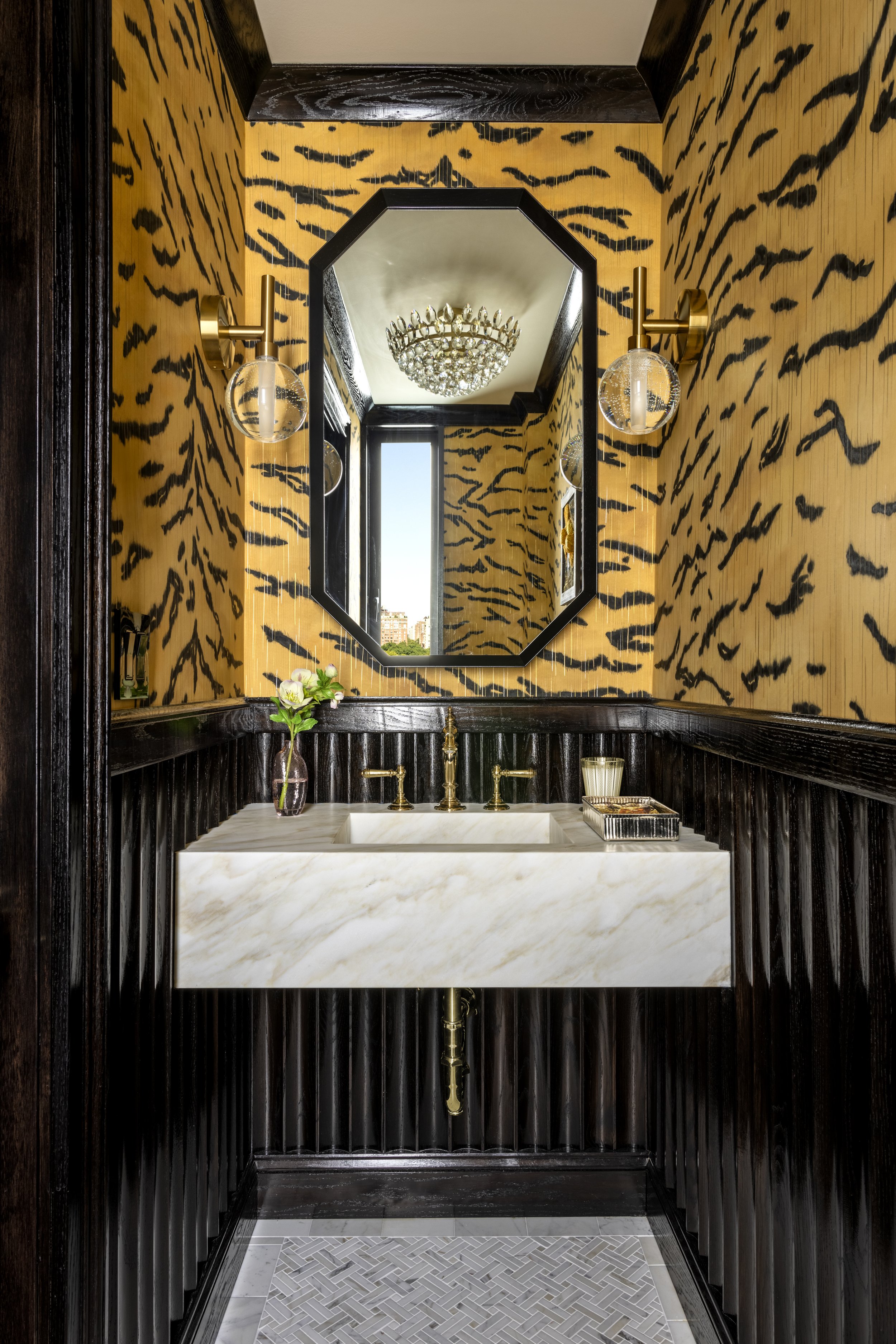 A luxurious bathroom with a marble sink, black wood paneling, yellow tiger stripe wallpaper, gold fixtures, a large octagonal mirror, a window showing a cityscape, and a chandelier reflected in the mirror.