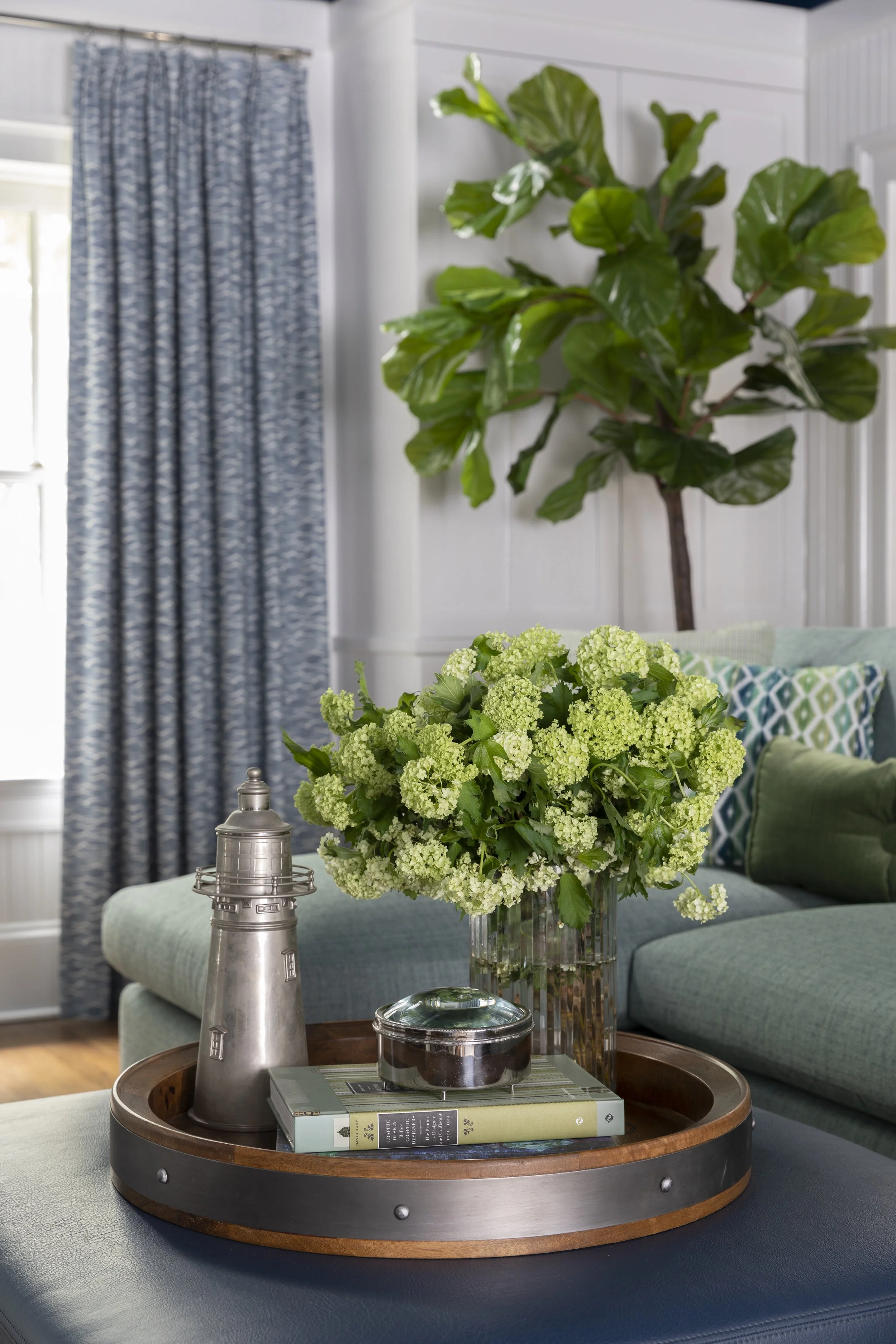 A living room with a round wooden table holding a glass vase with white-green hydrangeas, a metallic coffee grinder, a container, and books. In the background, a couch with patterned pillows, a window with patterned curtains, and a large indoor fiddl