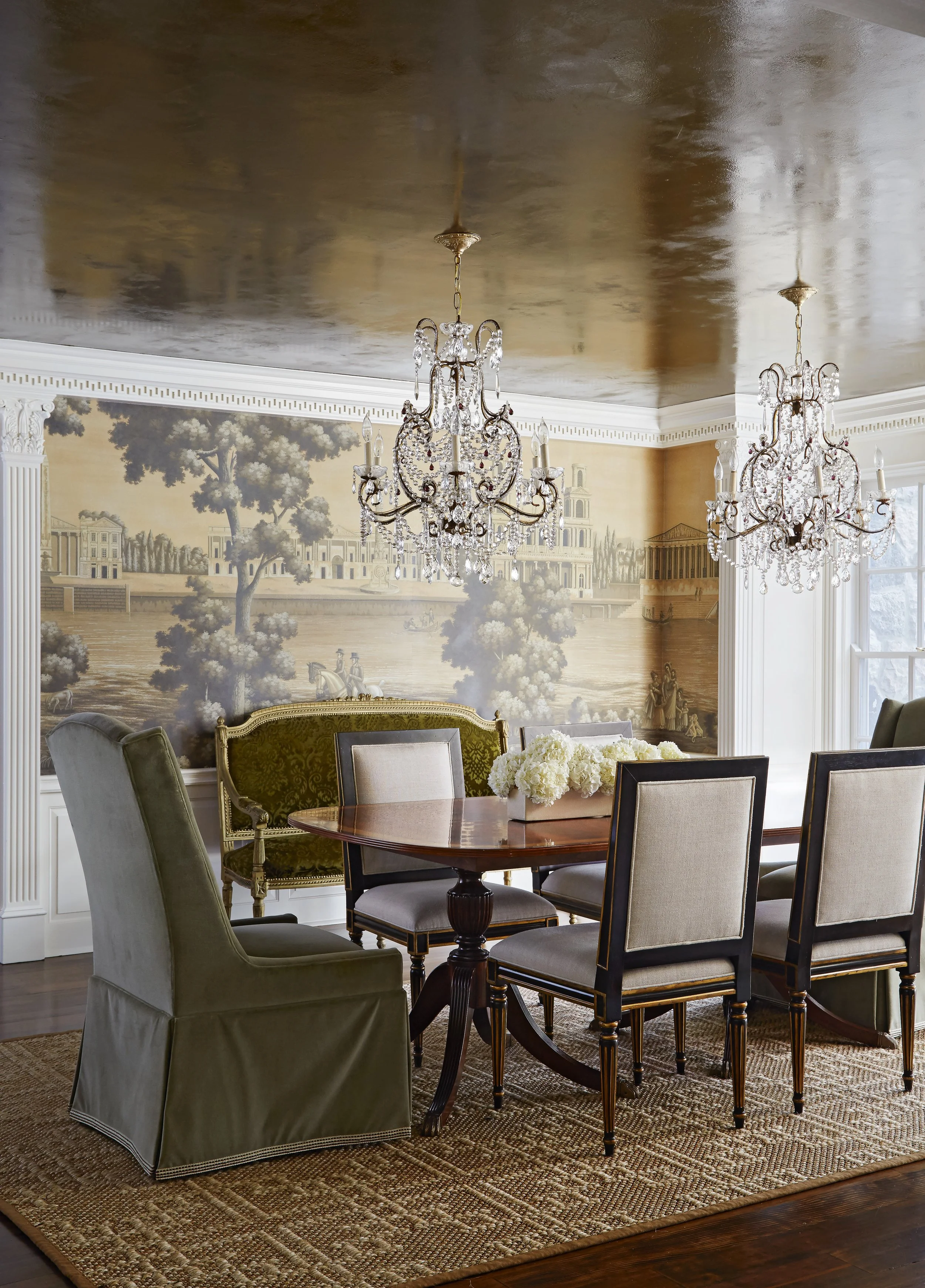 A dining room with a round wooden table, six upholstered chairs, a floral centerpiece, an ornate mural on the wall, and two large crystal chandeliers hanging from a gold ceiling.
