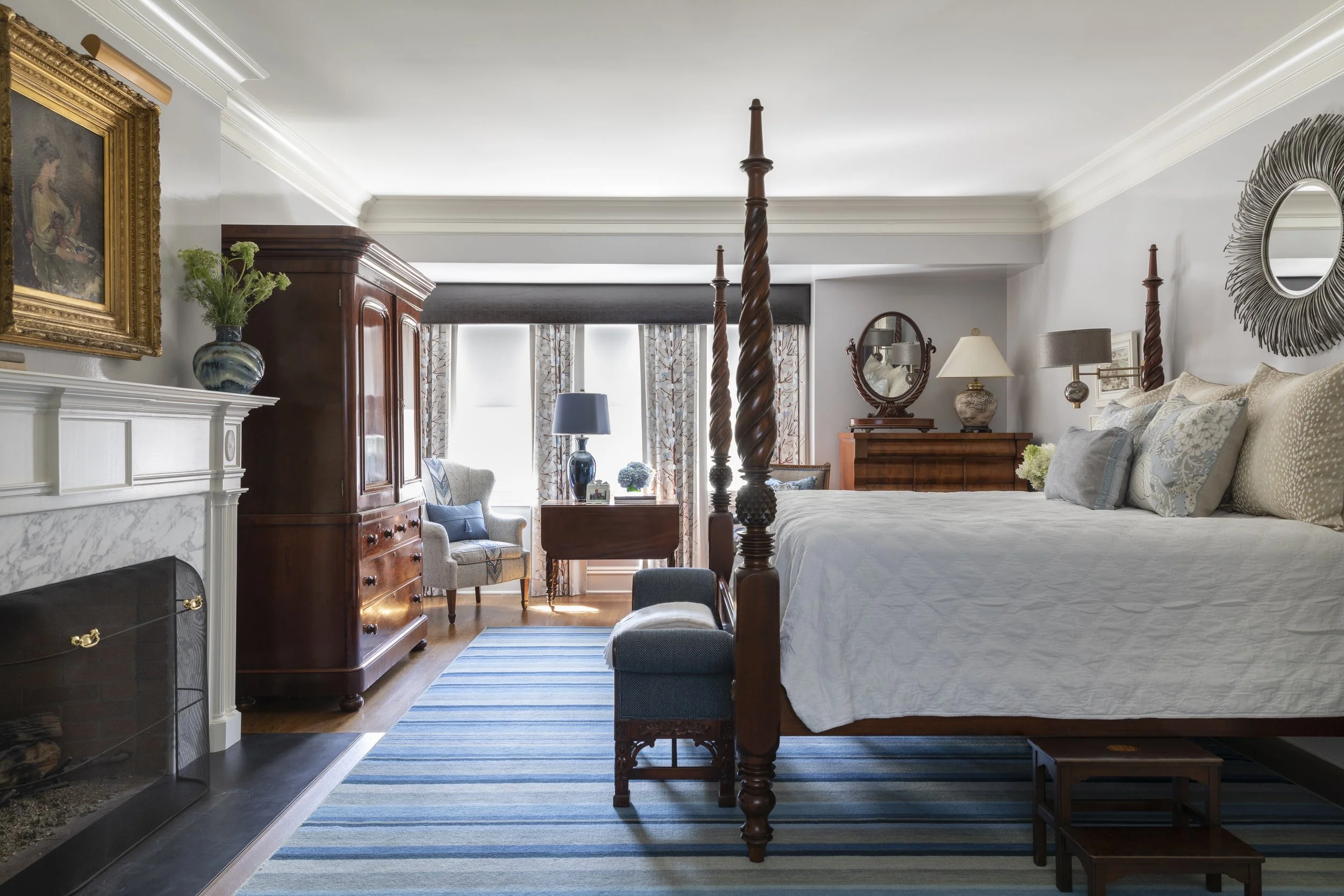 A bedroom with a four-poster wooden bed, a bedside table with a lamp, a large mirror on the wall, a wooden wardrobe, a fireplace with a marble mantel and a vase with flowers, and patterned curtains on the window. The room has white walls and a blue s