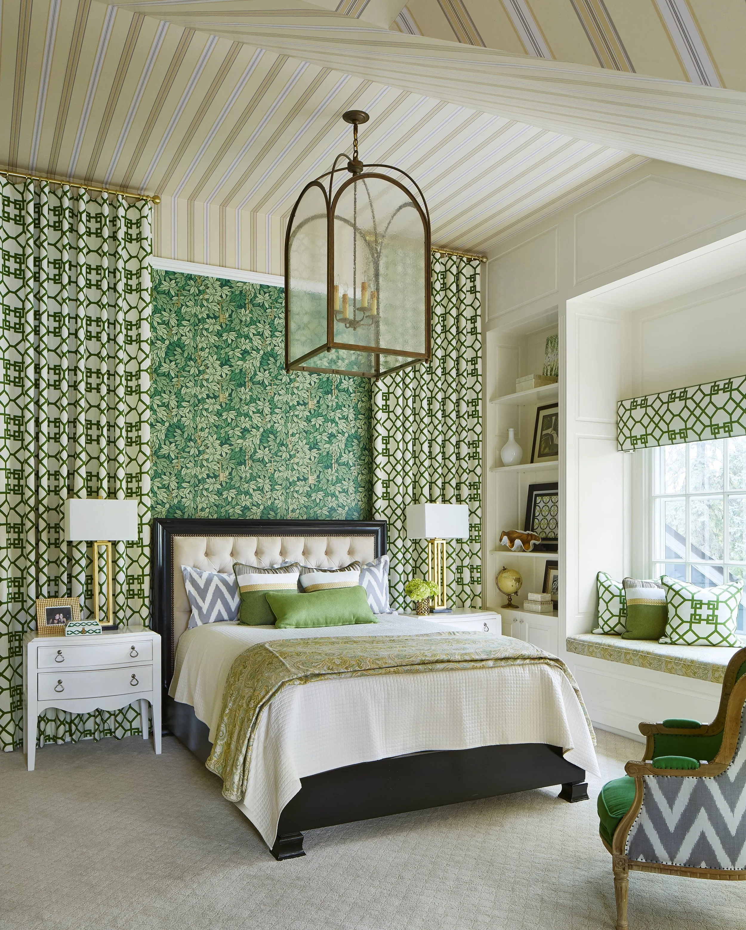 A bedroom with a white bed, green and gray pillows, and a decorative bedspread. There are nightstands with lamps, framed photos, and decorative objects. The room has patterned curtains and wall coverings in shades of green and cream, with a window se