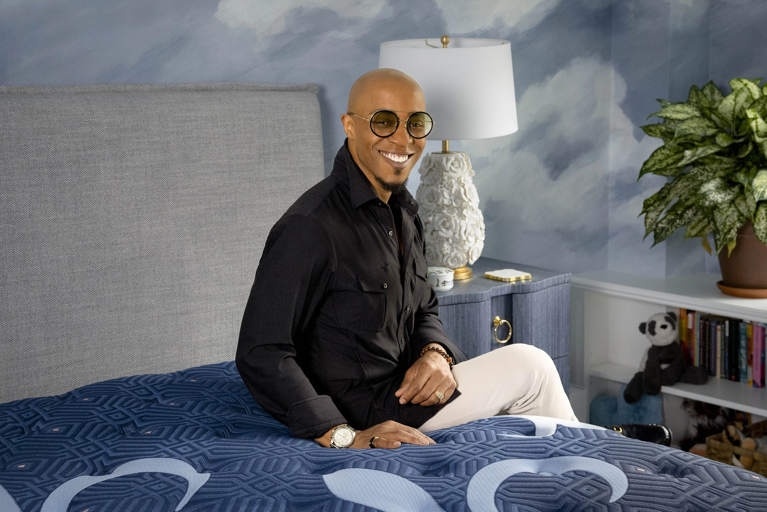 Corey Damen Jenkins with glasses and a beard, sitting on a bed in a room with blue walls, a white bedside lamp with a floral textured base, a large green plant, a bookshelf with stuffed animals, and books.