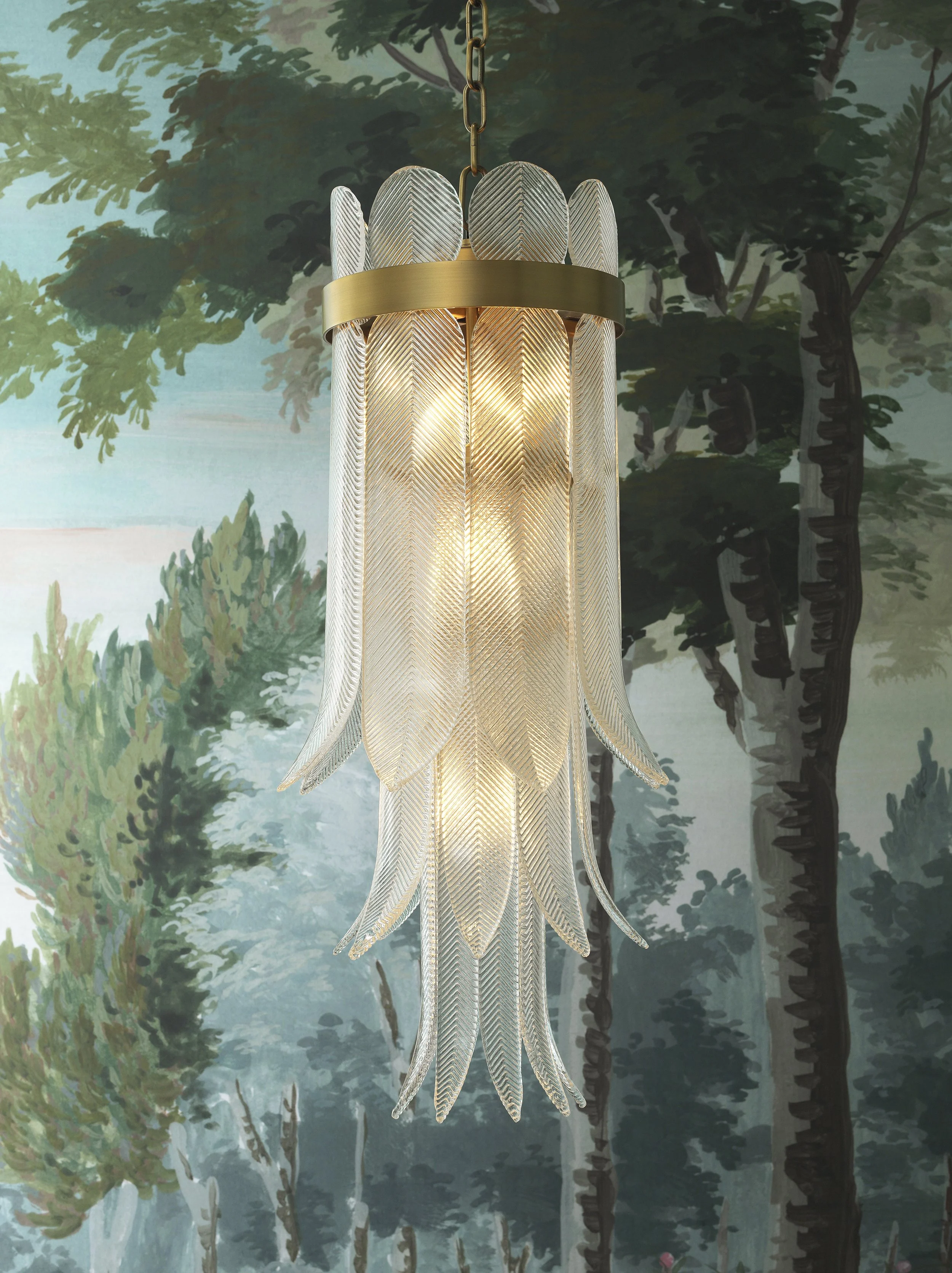 A chandelier with a gold band and tall, feather-like glass panels hangs in front of a mural depicting a forest scene with trees and foliage.