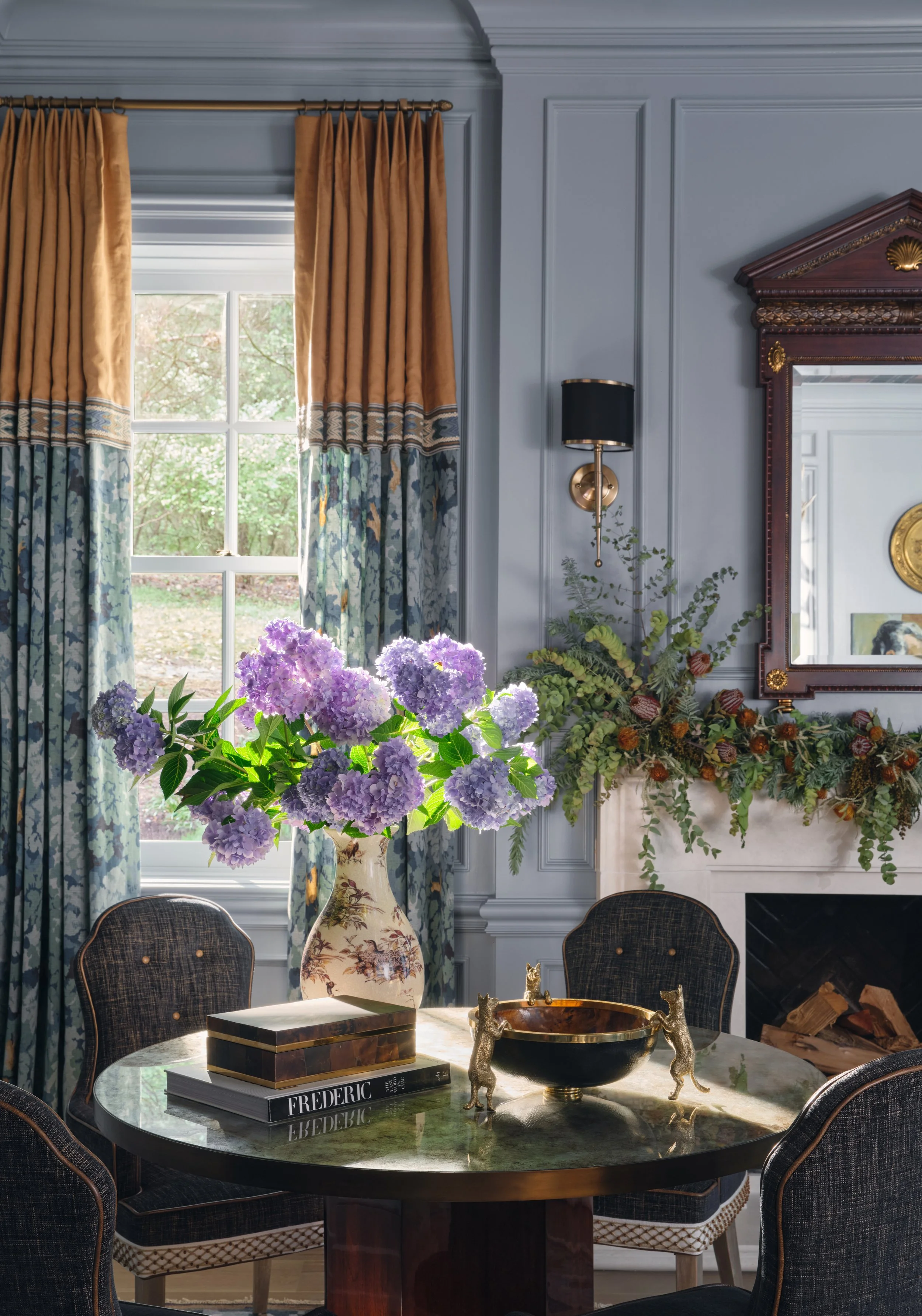 A dining room with a round marble table, surrounded by dark upholstered chairs, decorated with a large floral arrangement of purple hydrangeas in a beige vase, and gold decorative animals on the table. The room features a window with gold and blue cu
