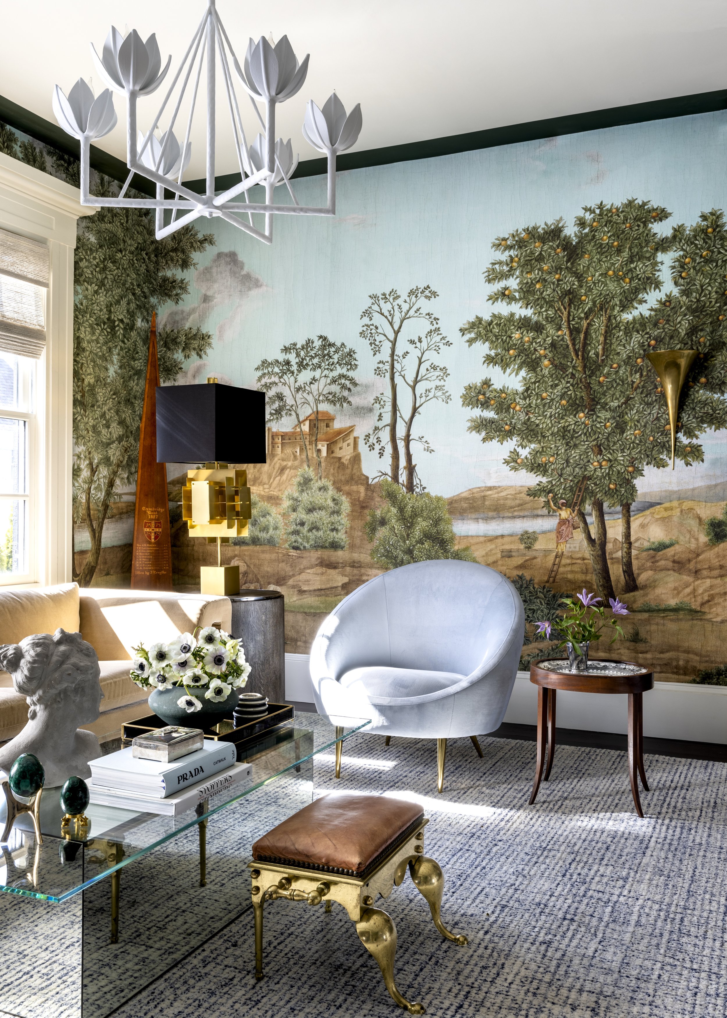 A stylish living room with a large painted mural of trees and a castle on the wall, a white curved armchair, a glass coffee table with books and decorative objects, a floral arrangement, a small side table with purple flowers, a modern chandelier, an