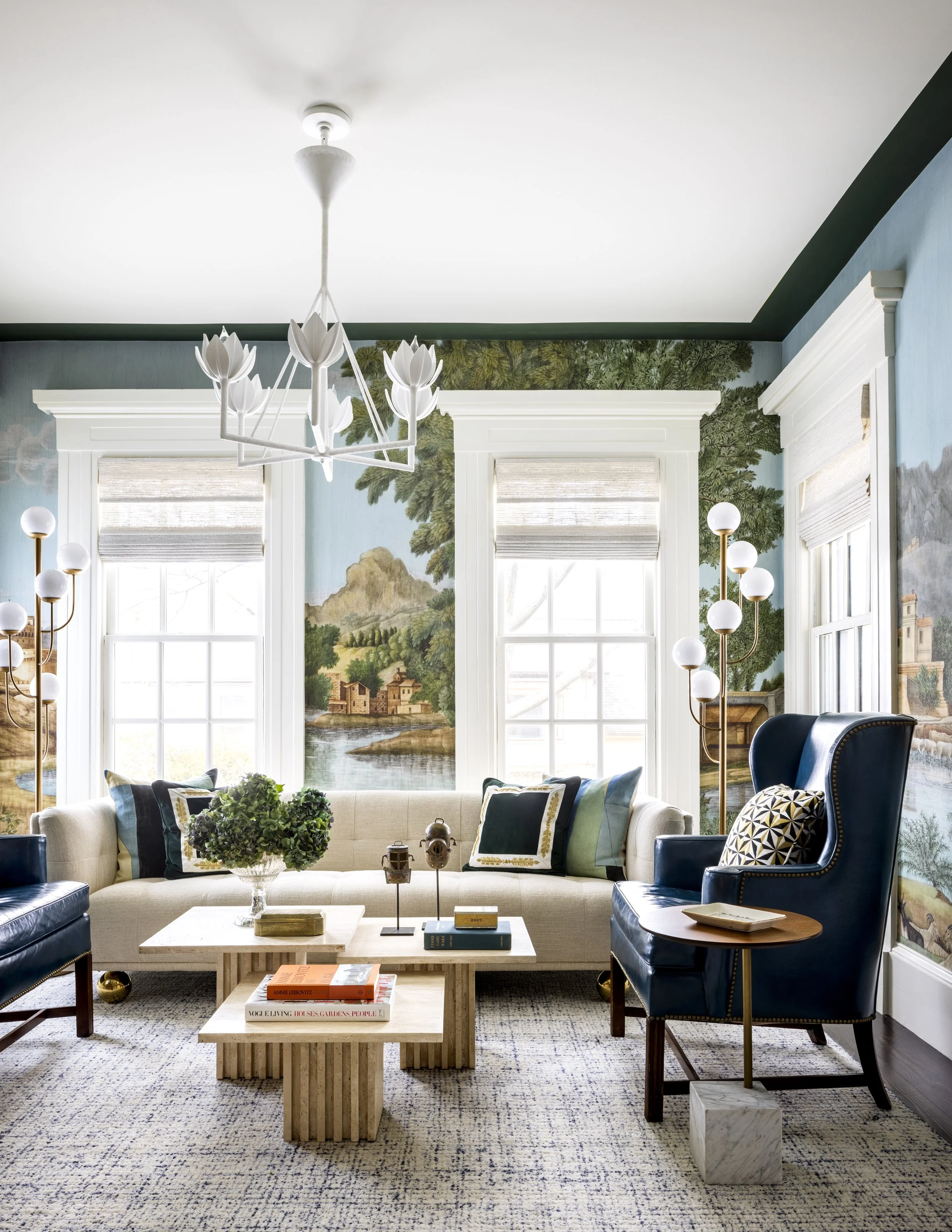 Living room with blue walls, large windows, painting of a landscape, beige sofa with pillows, black and blue armchairs, white coffee table with books, plant, and decor, white chandelier with tulip-shaped bulbs, various floor lamps, patterned rug.