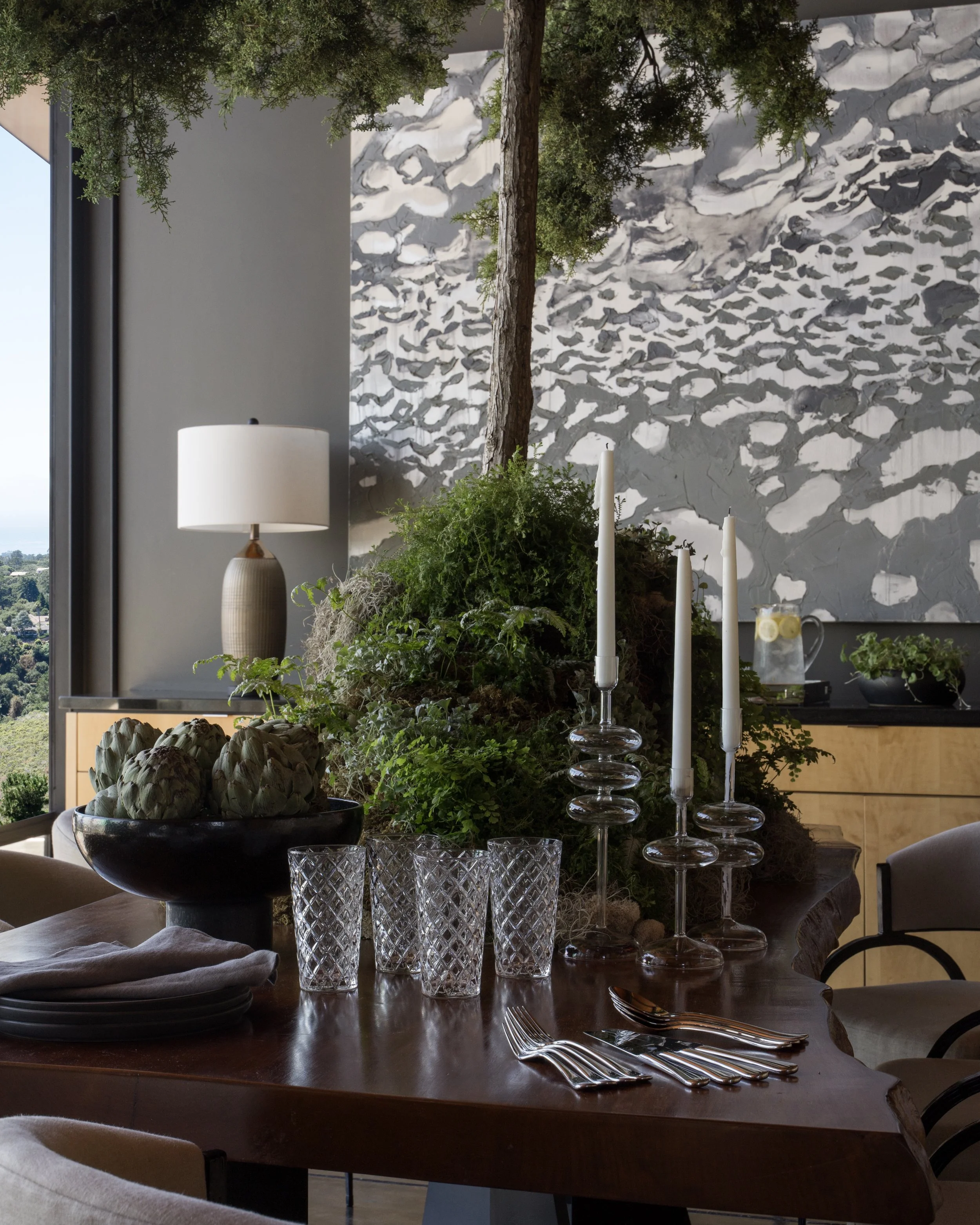 A dining table decorated with white candles, glassware, and greenery, with a modern abstract gray and white wall and a tree branch seen through a window in the background.