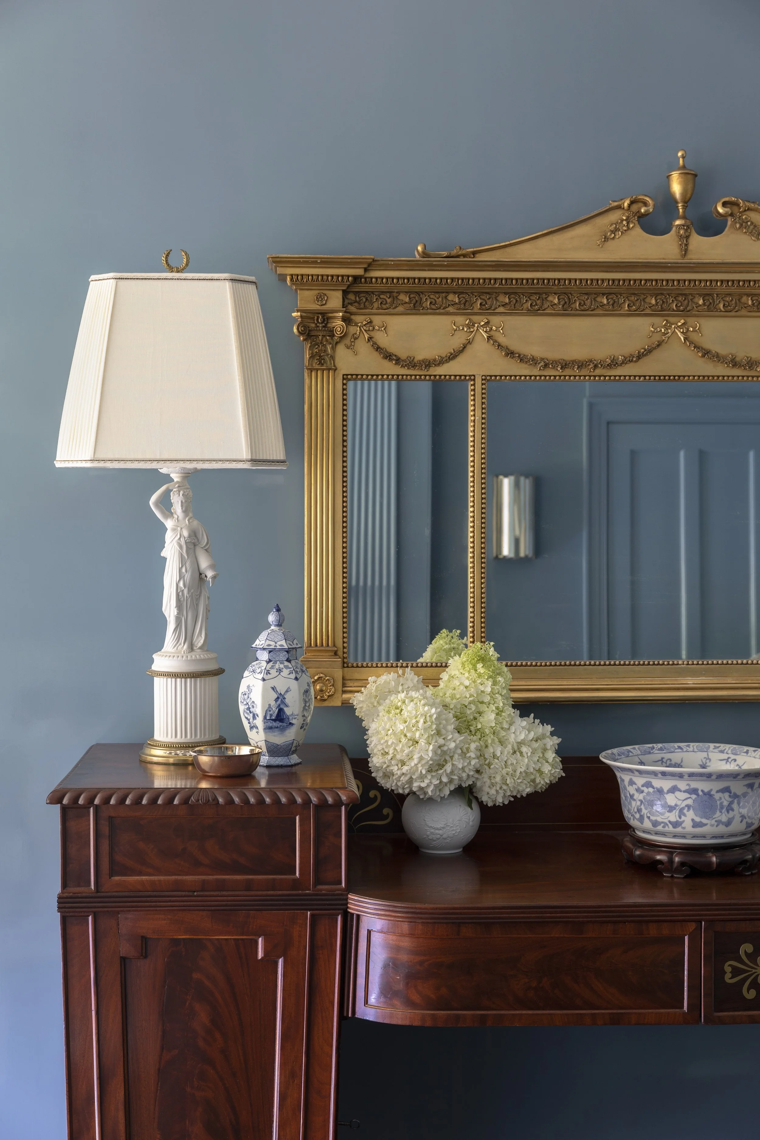 A wooden sideboard with a white lamp, a blue and white vase, a copper dish, white hydrangeas, and a blue and white porcelain bowl, set against a light blue wall with a gold-framed mirror and decorative elements.