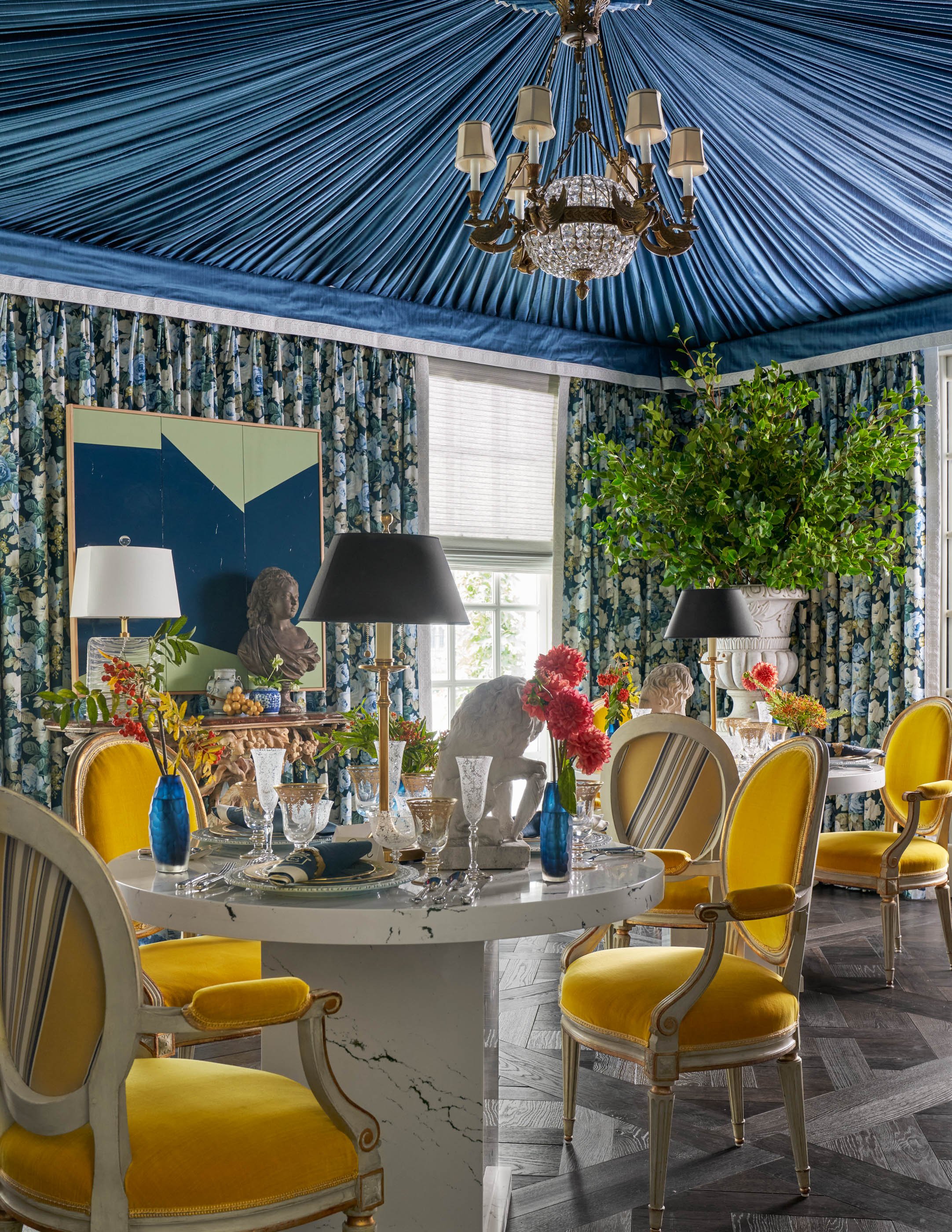 An elegant dining room with a blue fabric ceiling, floral curtains, a chandelier, and a large leafy plant. The table is set with glassware, vases with flowers, sculptures, and yellow upholstered chairs surrounding it.