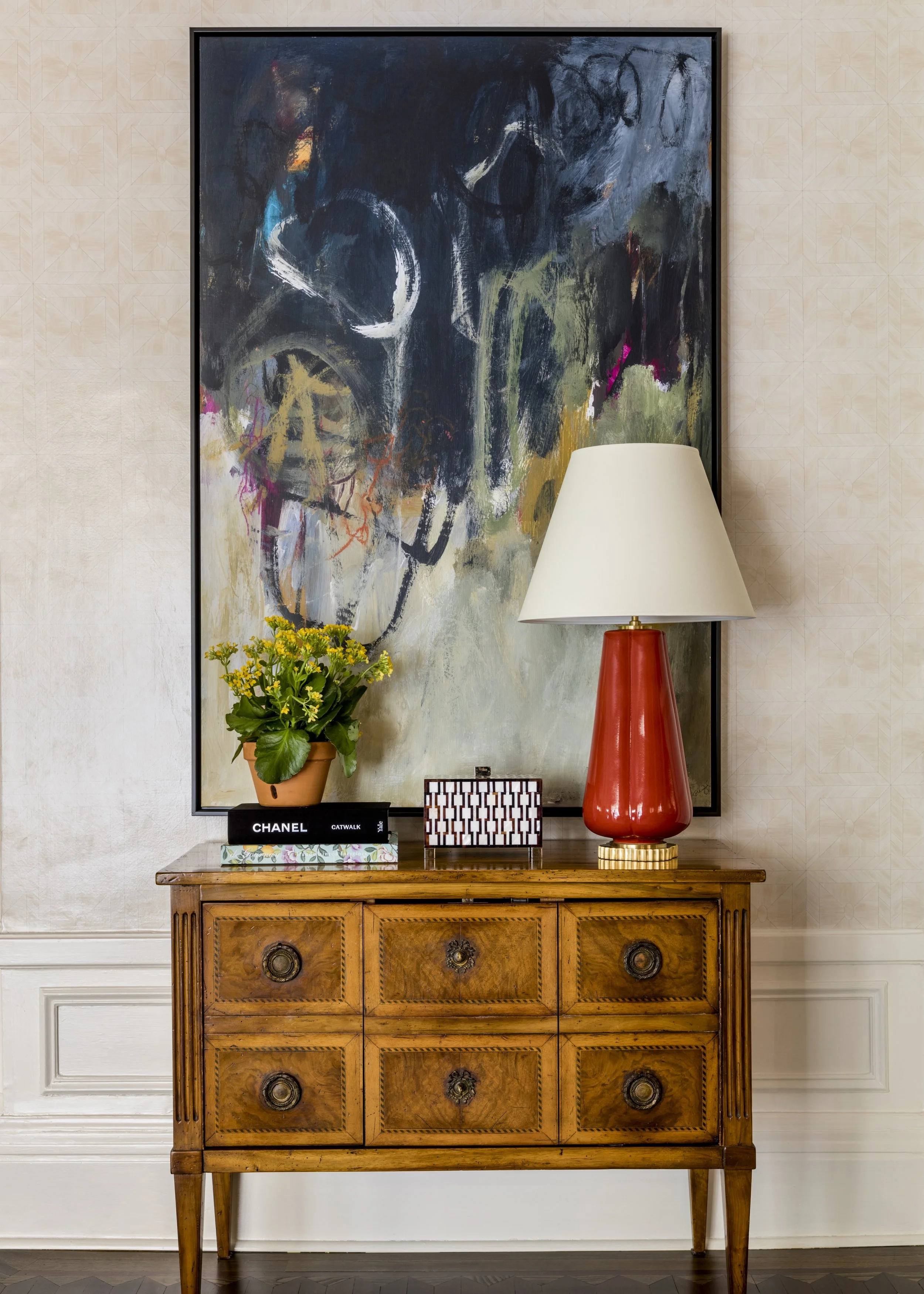 Interior decor featuring a wooden sideboard with various items on top, including books, a plant, a lamp, and decorative objects. Behind, a large abstract painting in dark colors, and the wall has a subtle patterned wallpaper.