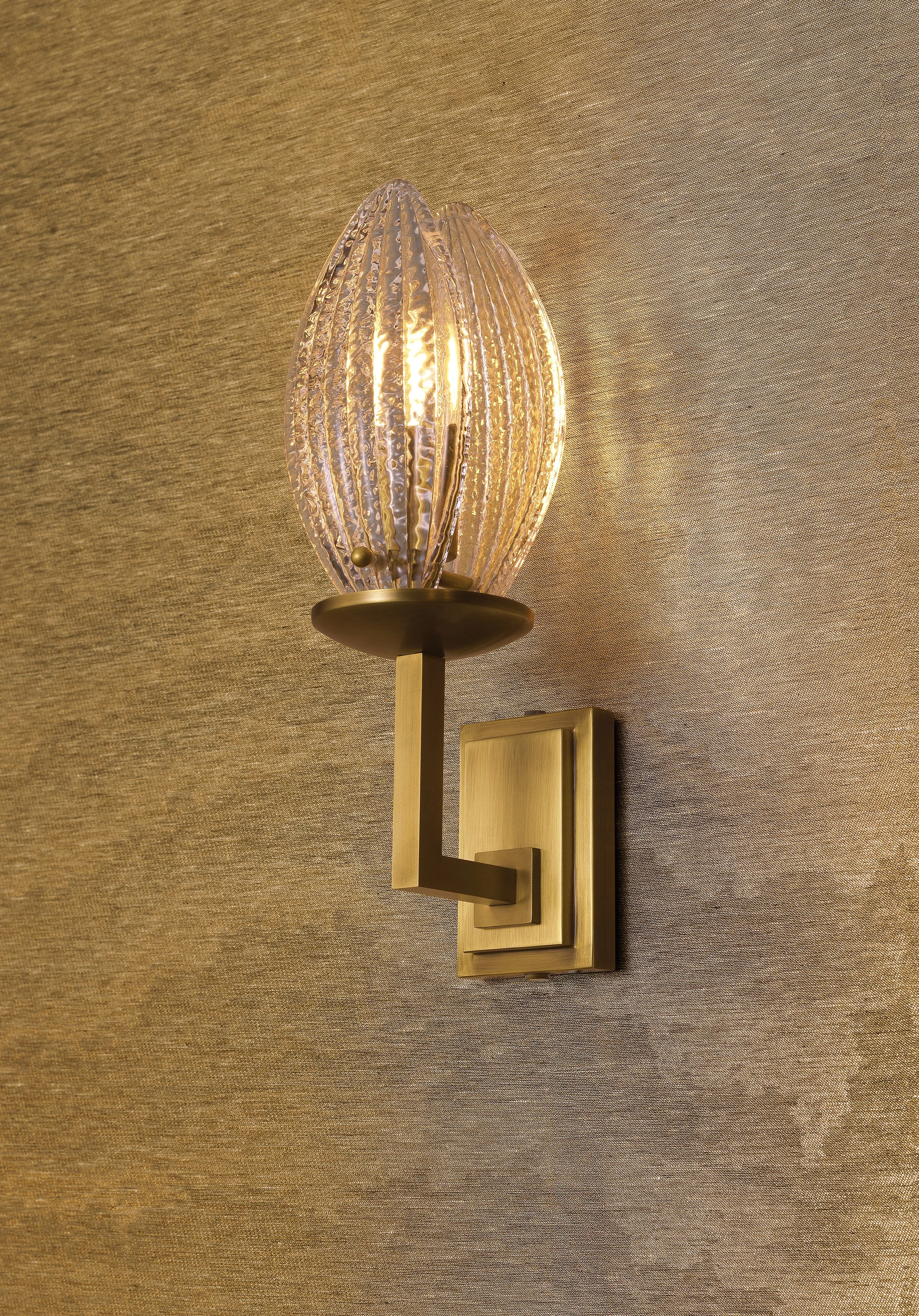 Wall-mounted light fixture with a gold base and textured glass shade emitting warm light, mounted on a gold wall plate.