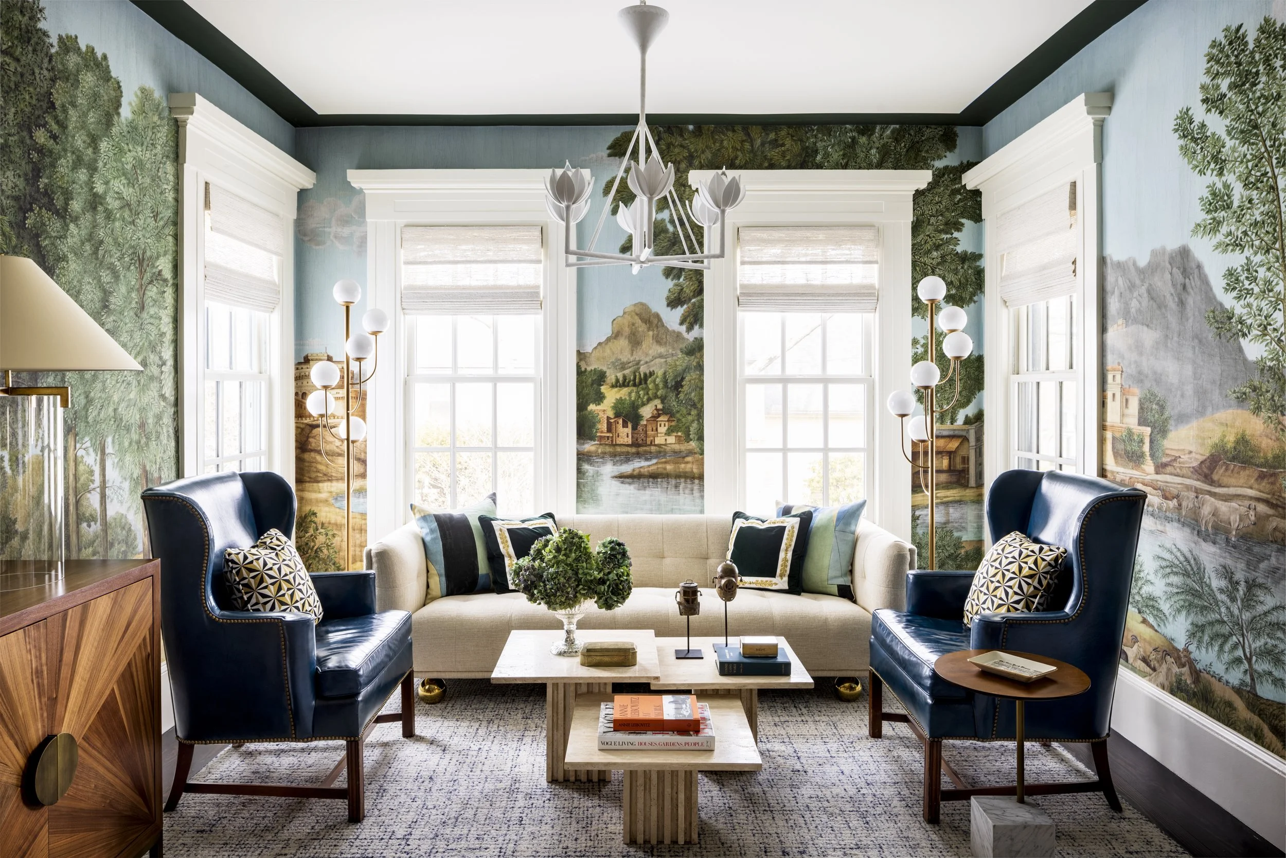 Living room with large mural window showing natural scenery, blue and white color scheme, modern chandelier, and furniture including a cream sofa, blue armchairs, and coffee table with books and decor.