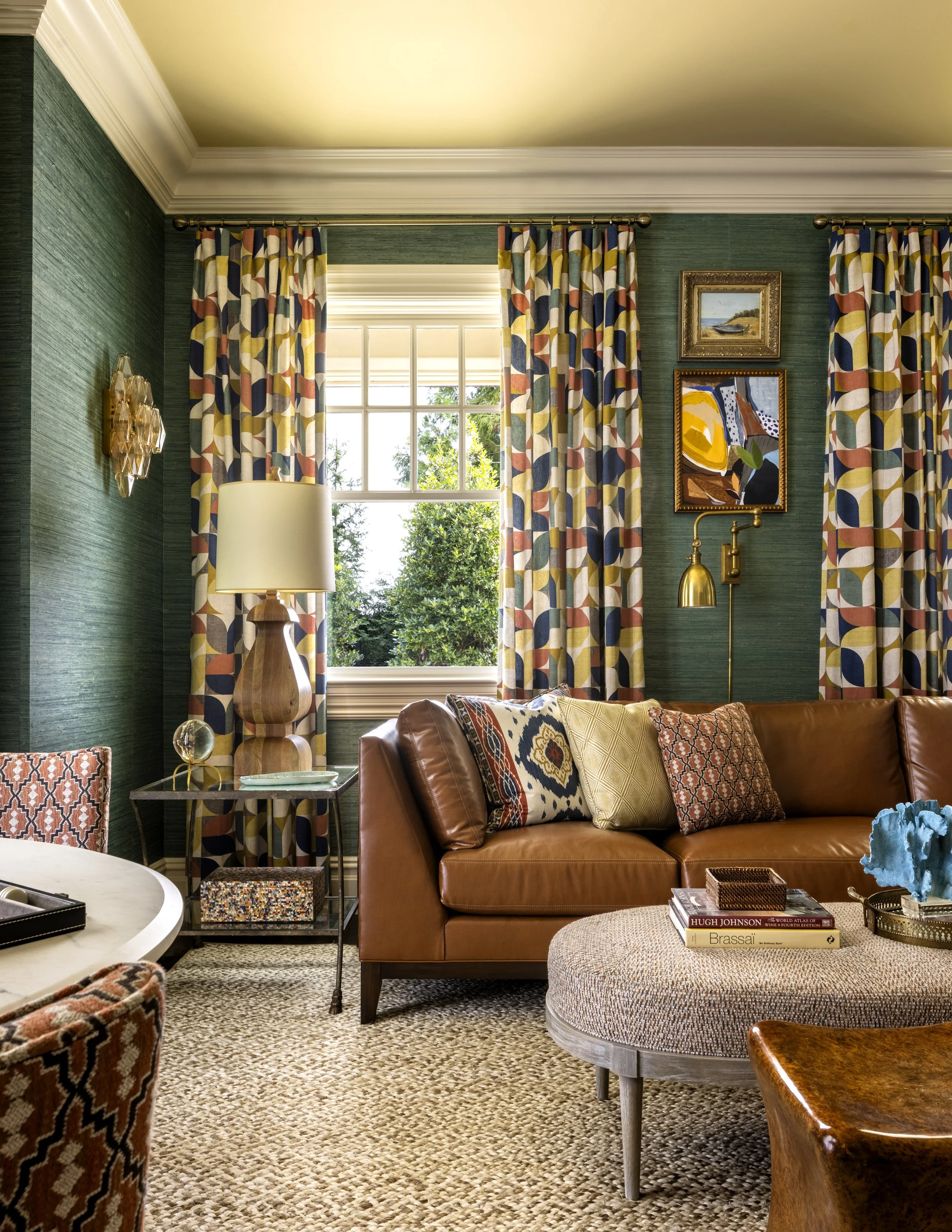 A cozy living room with large window, green textured walls, colorful patterned curtains, brown leather sofa with decorative pillows, side table with books, and artwork on the wall.