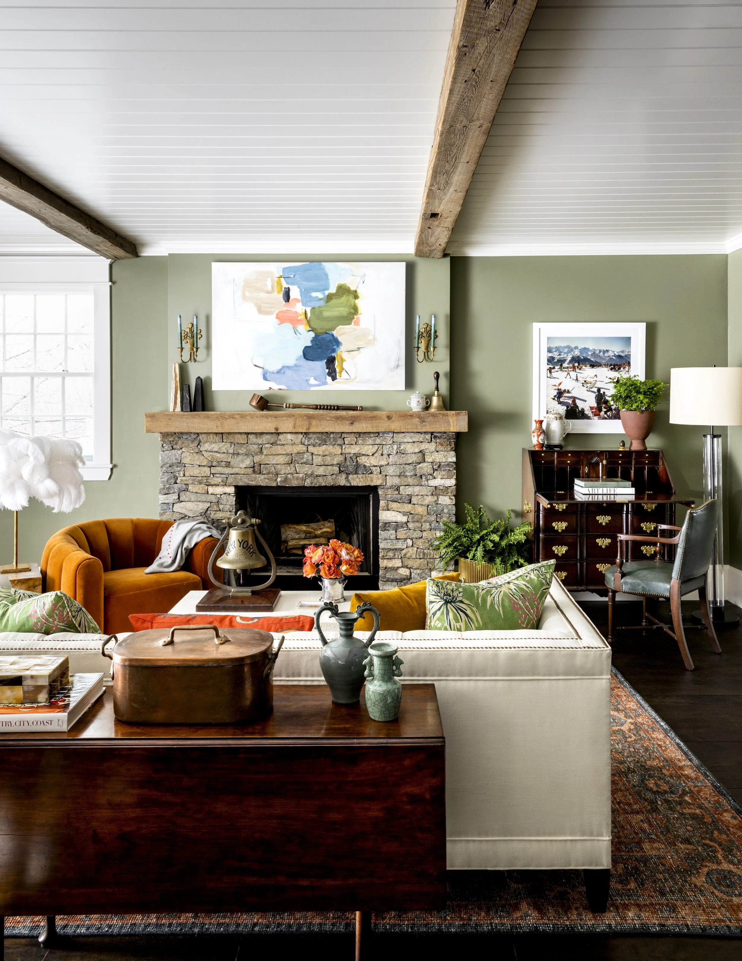 Living room with green walls, a stone fireplace with a wooden mantle, paintings, plants, and a variety of chairs and tables.