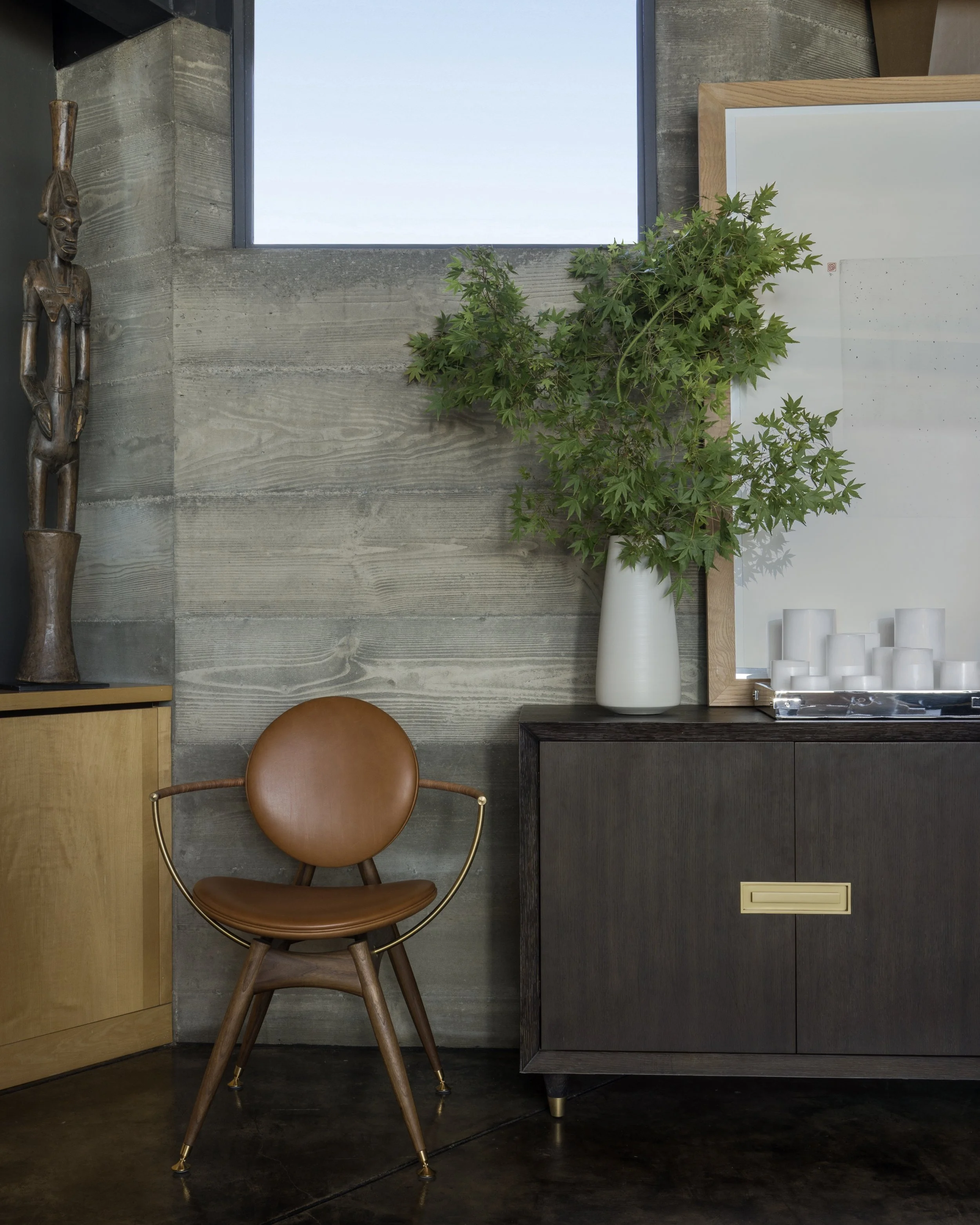 Interior decor with wood wall, a window, a wooden African sculpture, a white vase with green foliage, a framed art piece, a brown leather chair, and a dark cabinet.