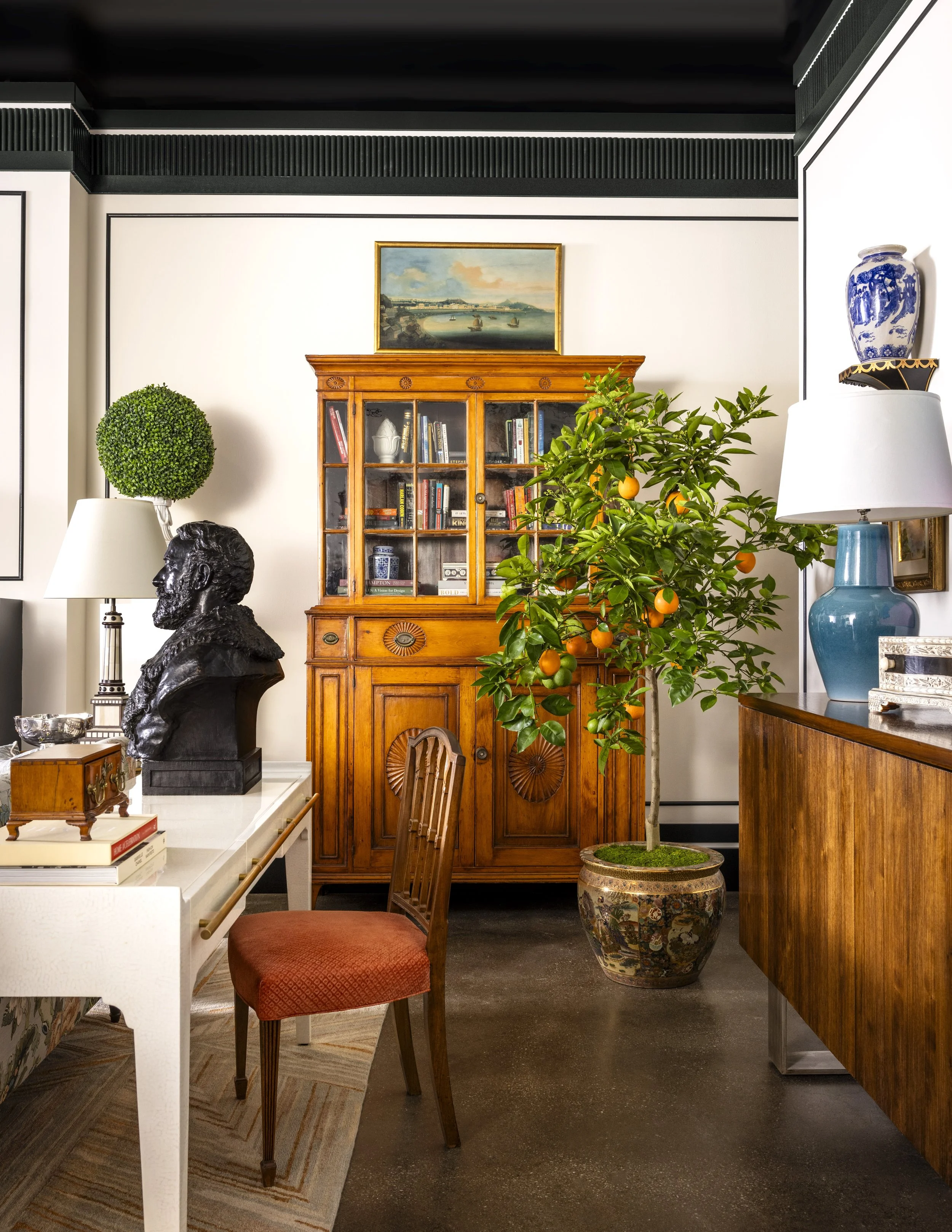 Interior of a room with a white wall, decorated with a landscape painting, a wooden cabinet with glass doors filled with books, a bust sculpture on a white table, and a potted orange tree. There are various lamps and decorative items, including a blu