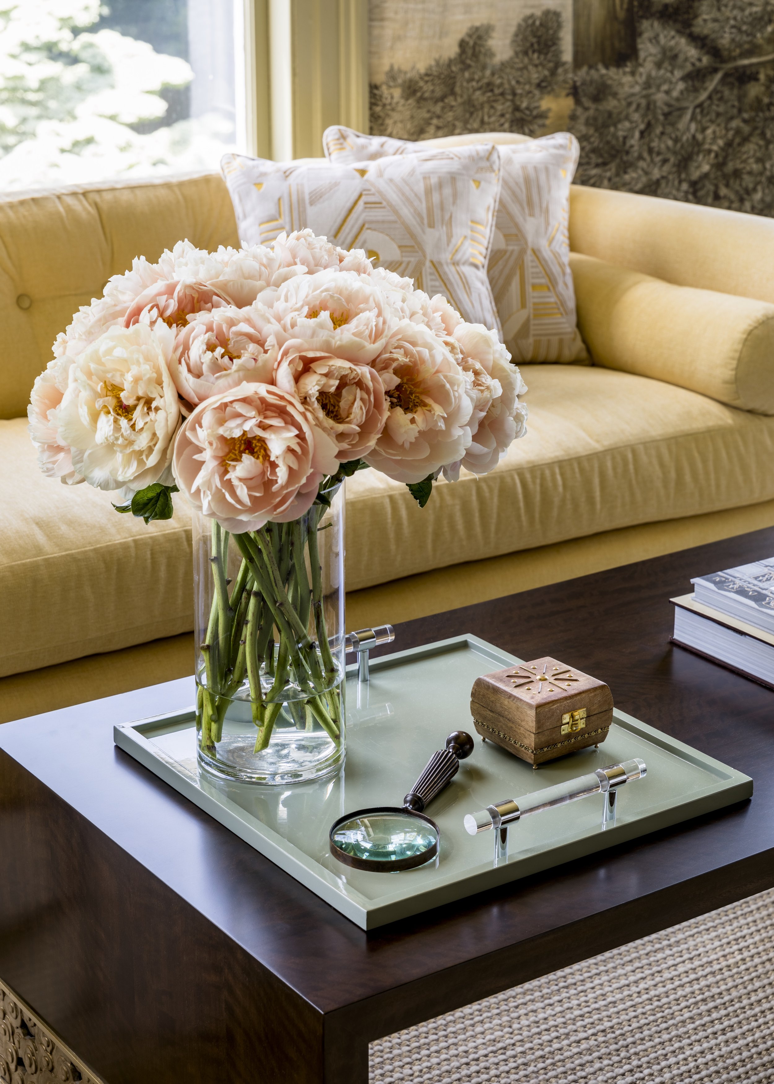 A living room with a yellow couch, decorative pillows, a window with outdoor greenery, a coffee table with a floral vase, books, and decorative items.