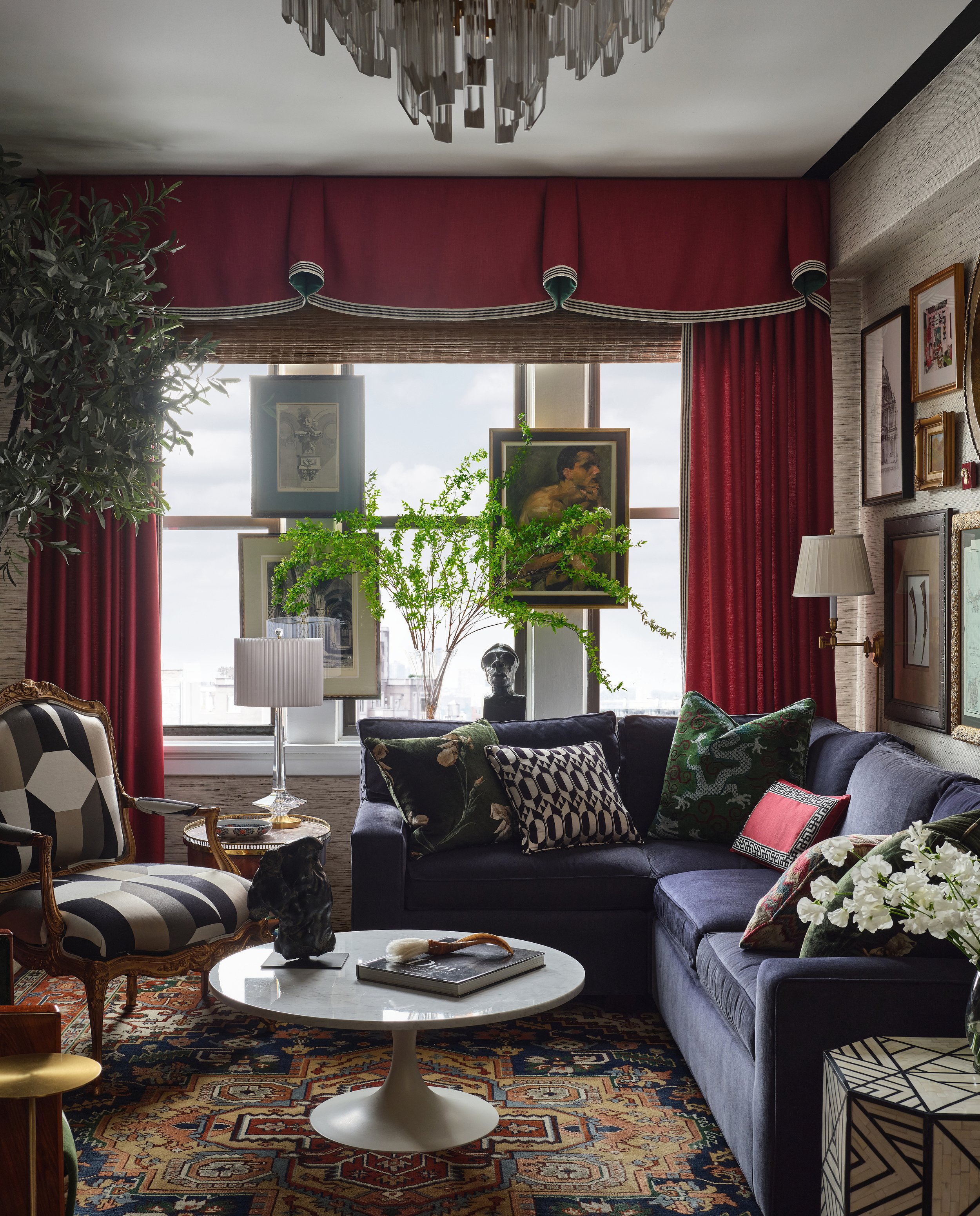 Living room with large window, red drapes, green plants, framed artwork, dark blue sofa with patterned cushions, and a white marble coffee table with a black bust and a book.