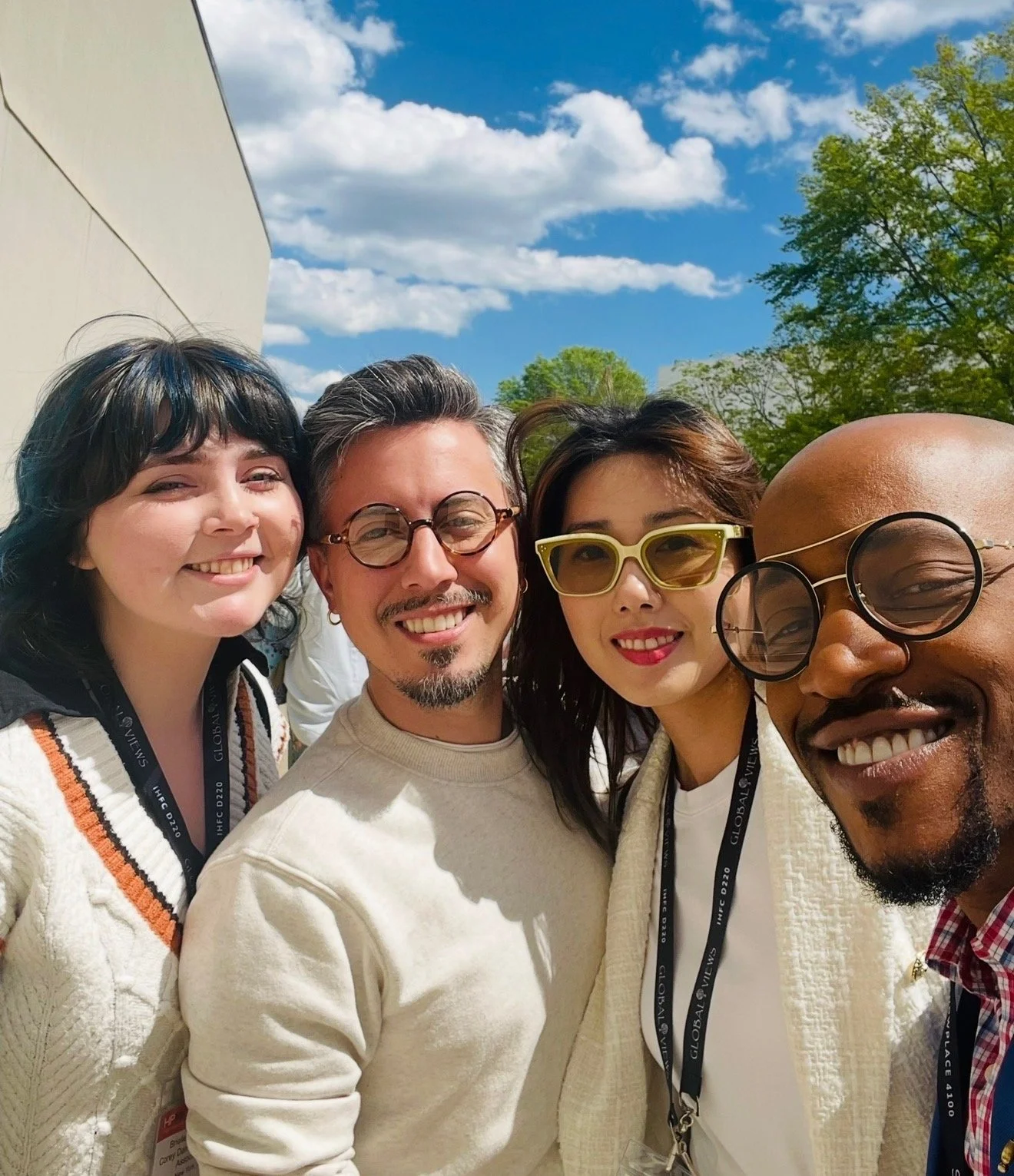 Group of five friends taking a selfie outdoors on a sunny day, smiling, with blue sky and green trees in the background.