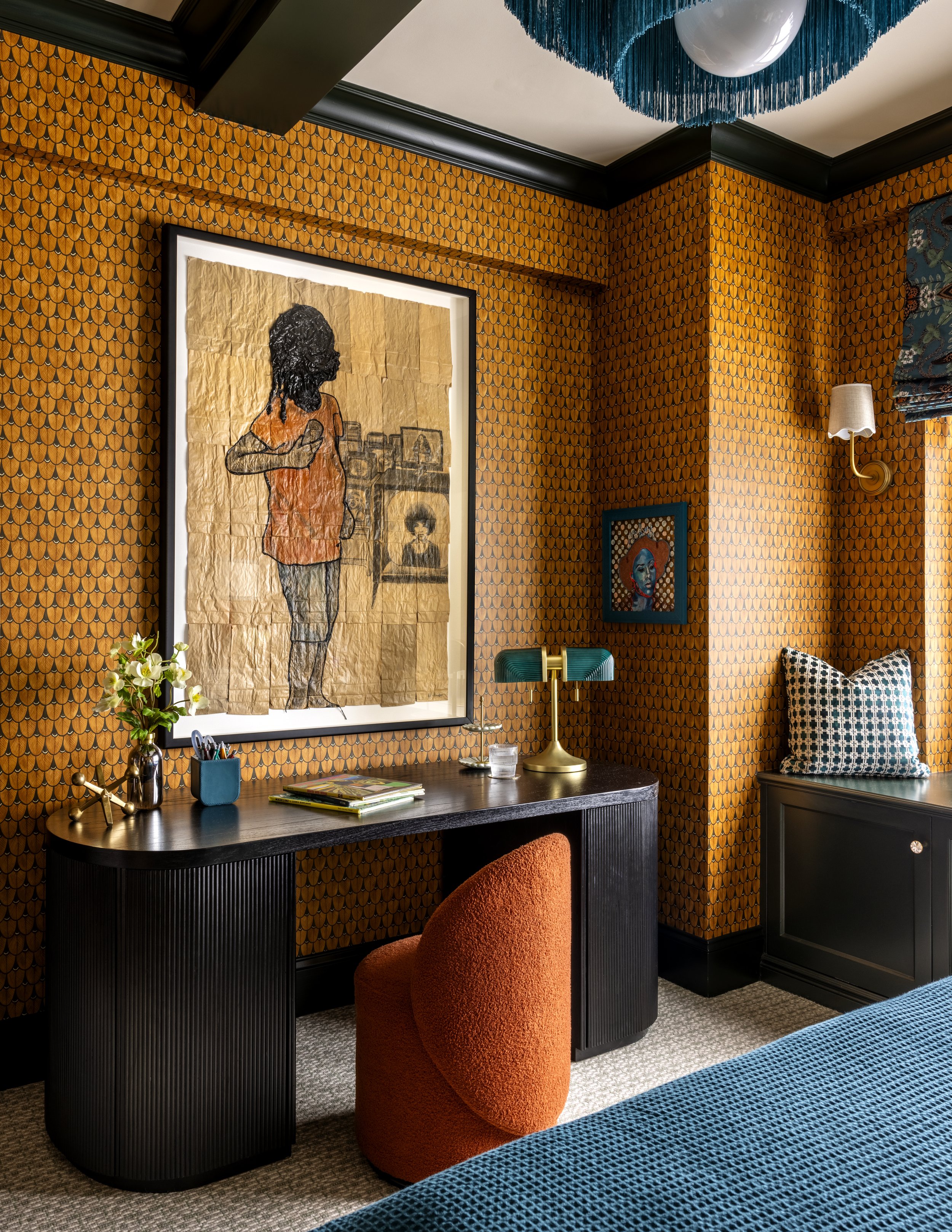 A cozy home office or reading nook with orange patterned wallpaper, a black desk with a curved edge, an orange plush chair, a brass and green desk lamp, and framed artwork on the wall. There is a small table with books and a vase of flowers, and a be
