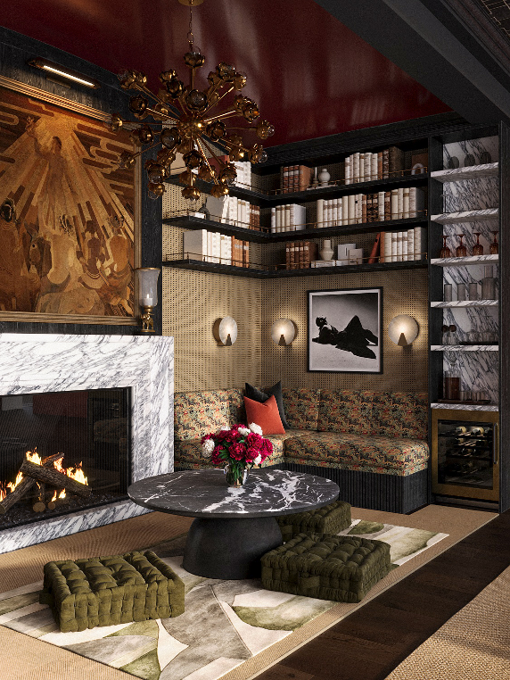 A cozy living room with a marble fireplace, a round black coffee table, green cushioned floor seats, a floral-patterned sofa with red and black pillows, and wall-mounted lights. Shelves with books and decorative items, a framed black and white photo,