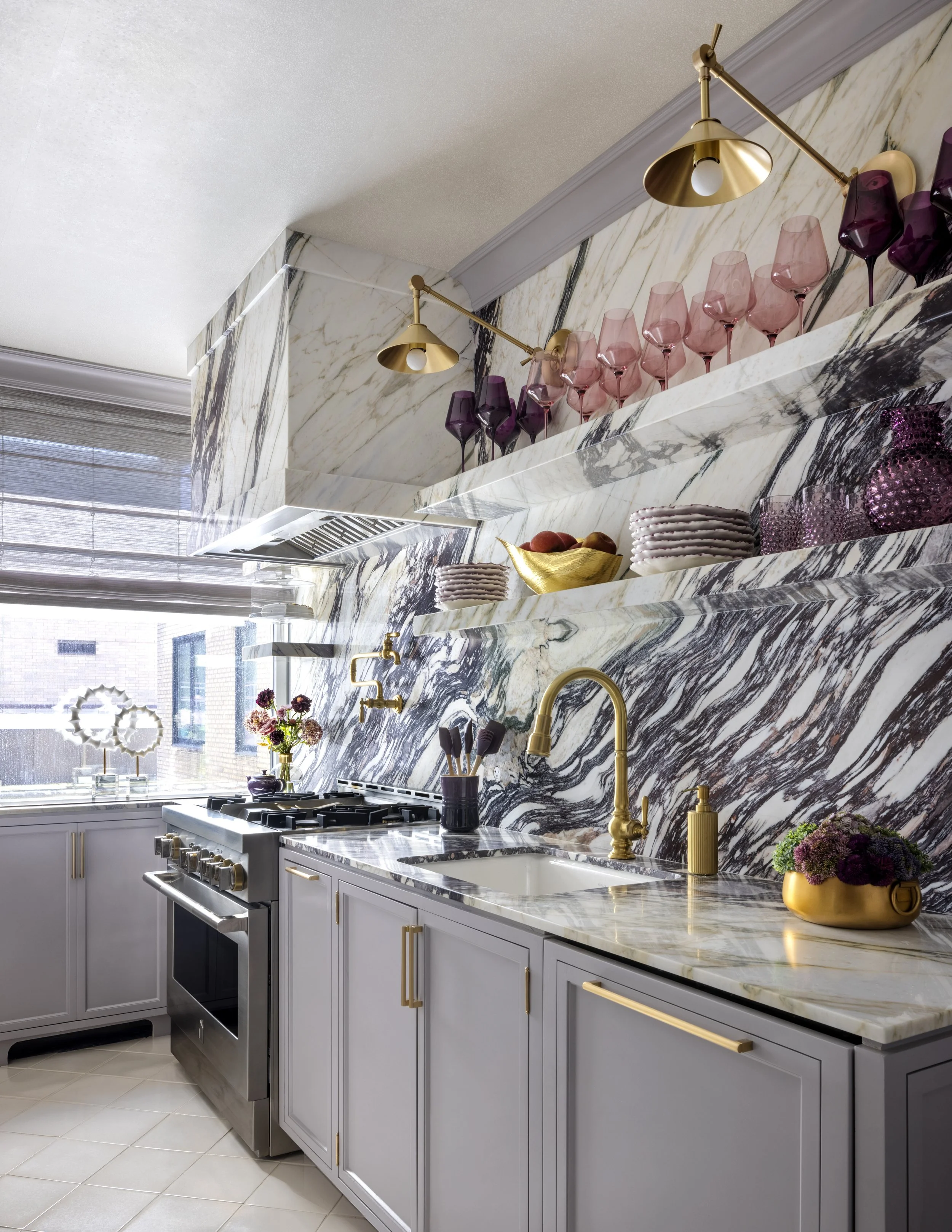Modern kitchen with marble countertops and backsplash, gold fixtures, purple and pink wine glasses on shelves, white cabinets with gold handles, and stainless steel stove.