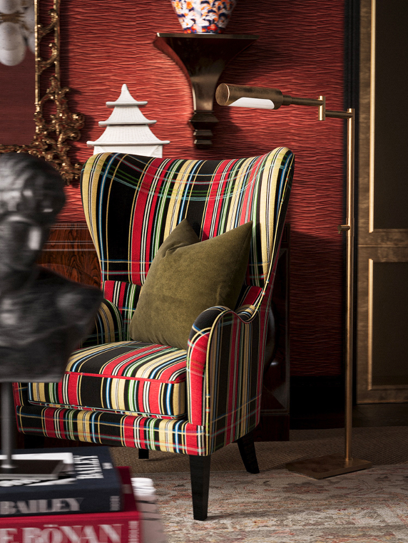 A colorful upholstered armchair with vertical stripes in red, green, yellow, and black, with a green pillow on it, placed in front of a red textured wall with decorative objects and a lamp.
